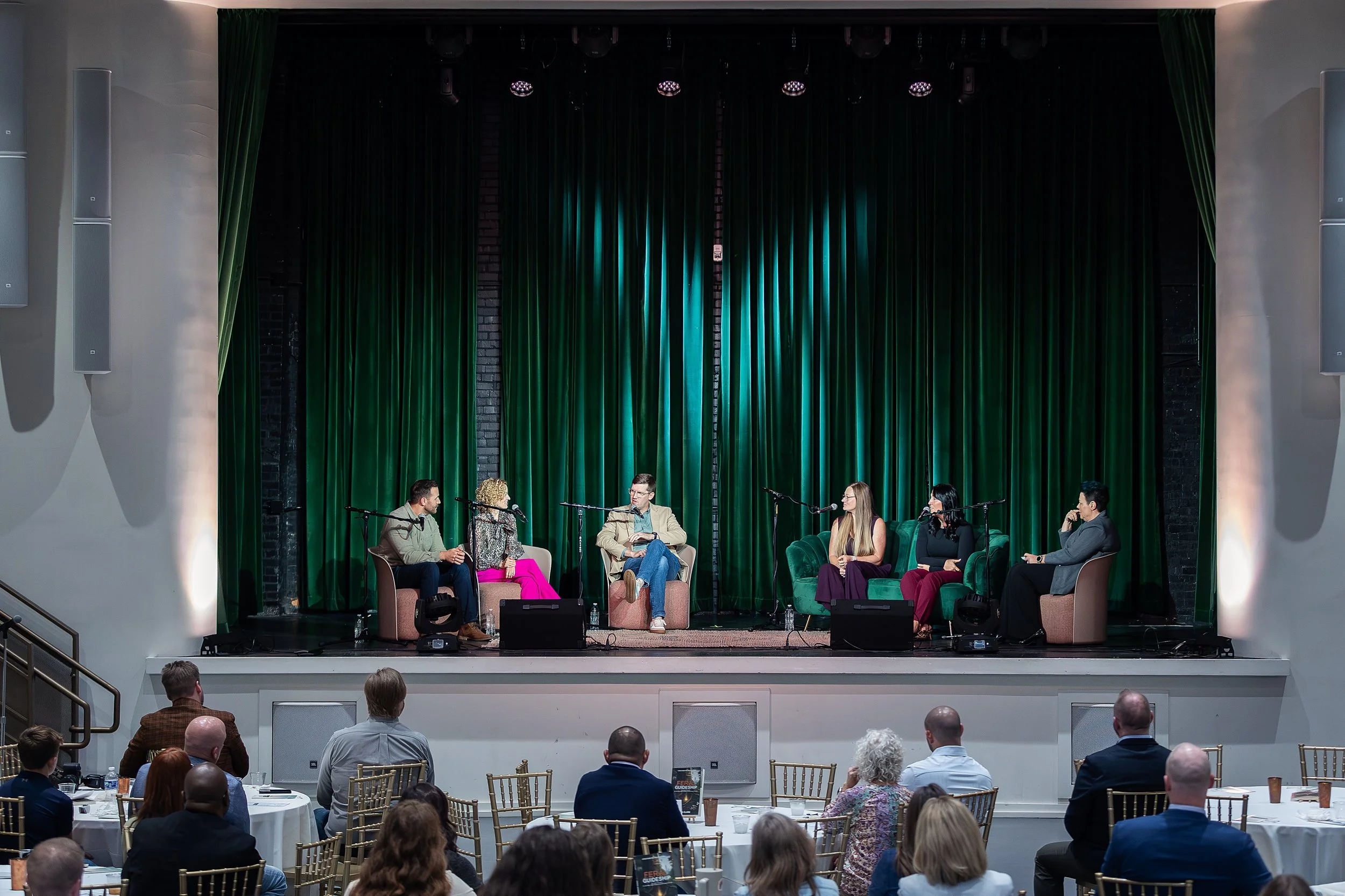 A panel discussion or interview taking place on a stage with six people seated in chairs. The stage has green curtains, and there is an audience seated at tables facing the stage.