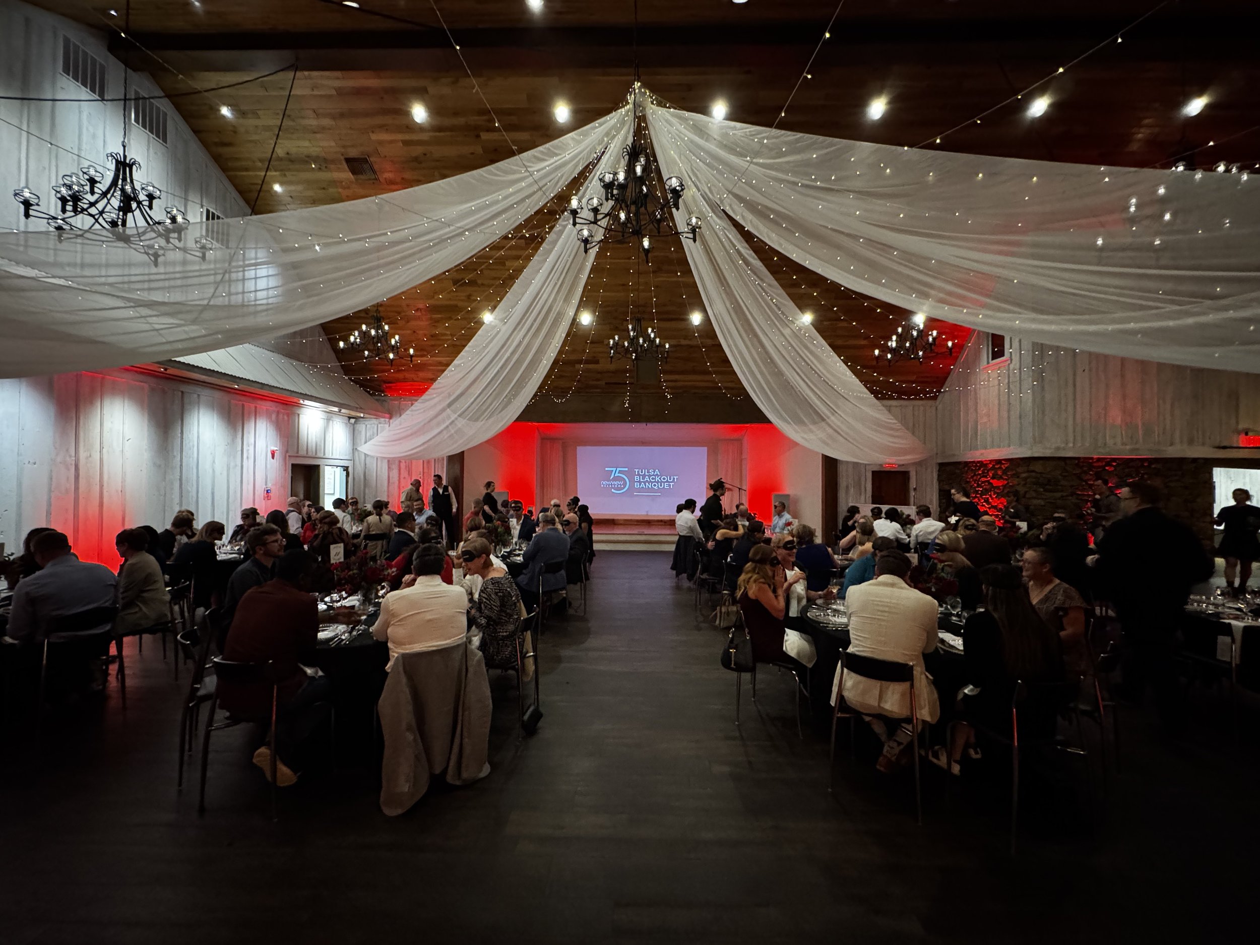An indoor banquet event with guests seated at round tables, decorated with flowers. The ceiling is draped with white fabric and string lights, and black chandeliers hang from the ceiling. The stage at the front displays a screen with the logo and tex