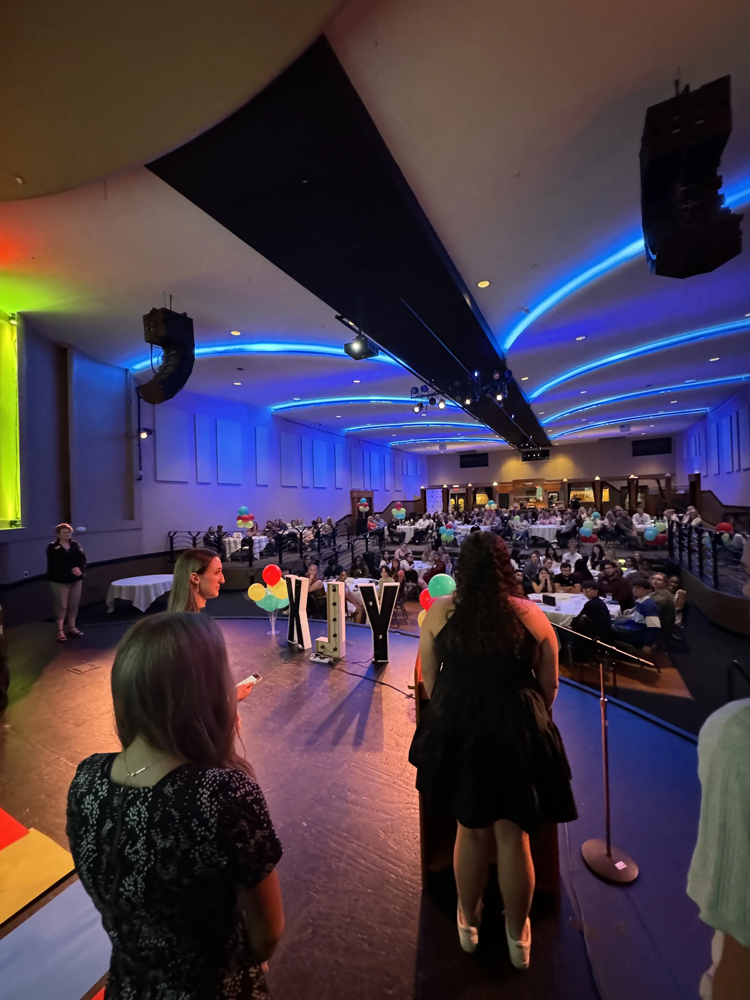 People gathered in a decorated banquet hall with blue lighting, balloons, and tables, attending an event or celebration.