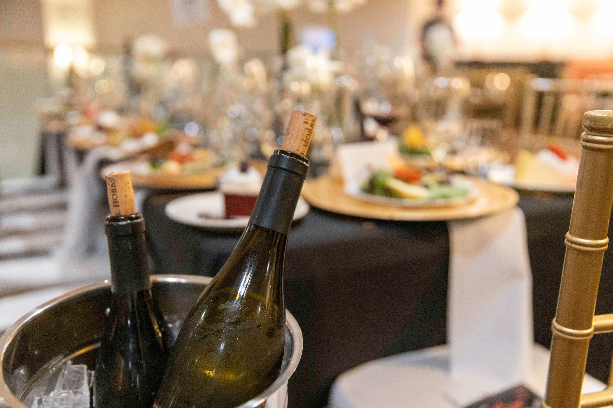 Two wine bottles submerged in an ice bucket on a table at a dining event, with a blurred background of plates and glasses.
