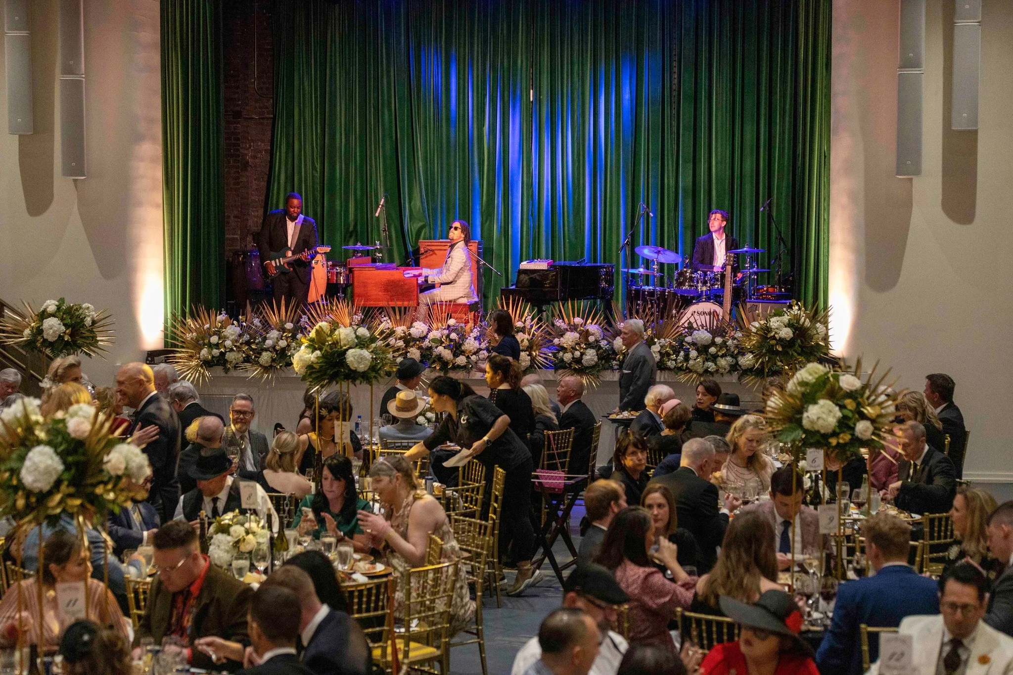 An indoor event with a large crowd seated at decorated tables, a stage with a band performing including a pianist in a gray suit, a guitarist, and a drummer, with a green curtain backdrop and floral arrangements in the foreground.