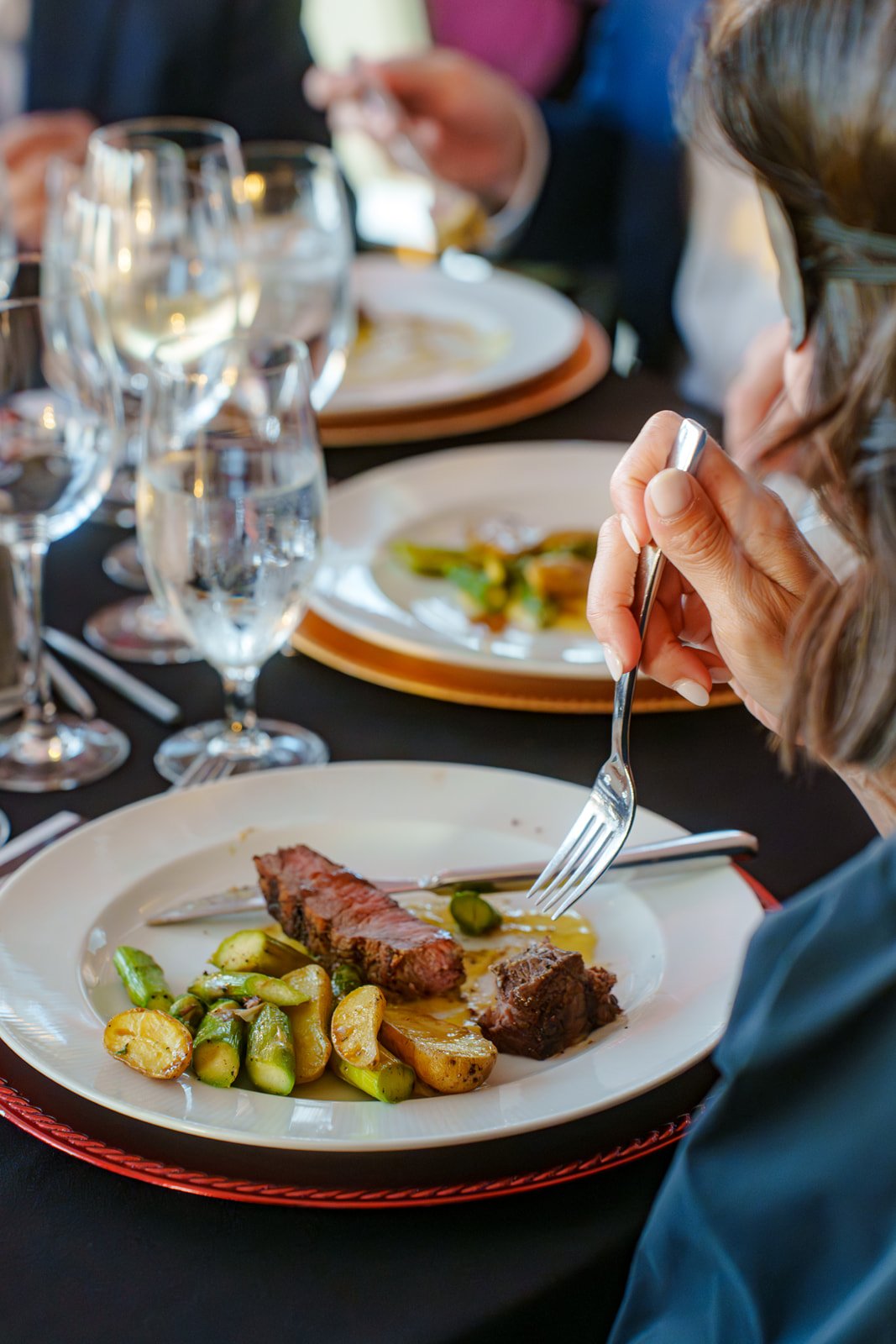 Close-up of a person eating at a fine dining restaurant, with a plate of steak, potatoes, and green beans in front of them, and a table set with wine glasses and plates.