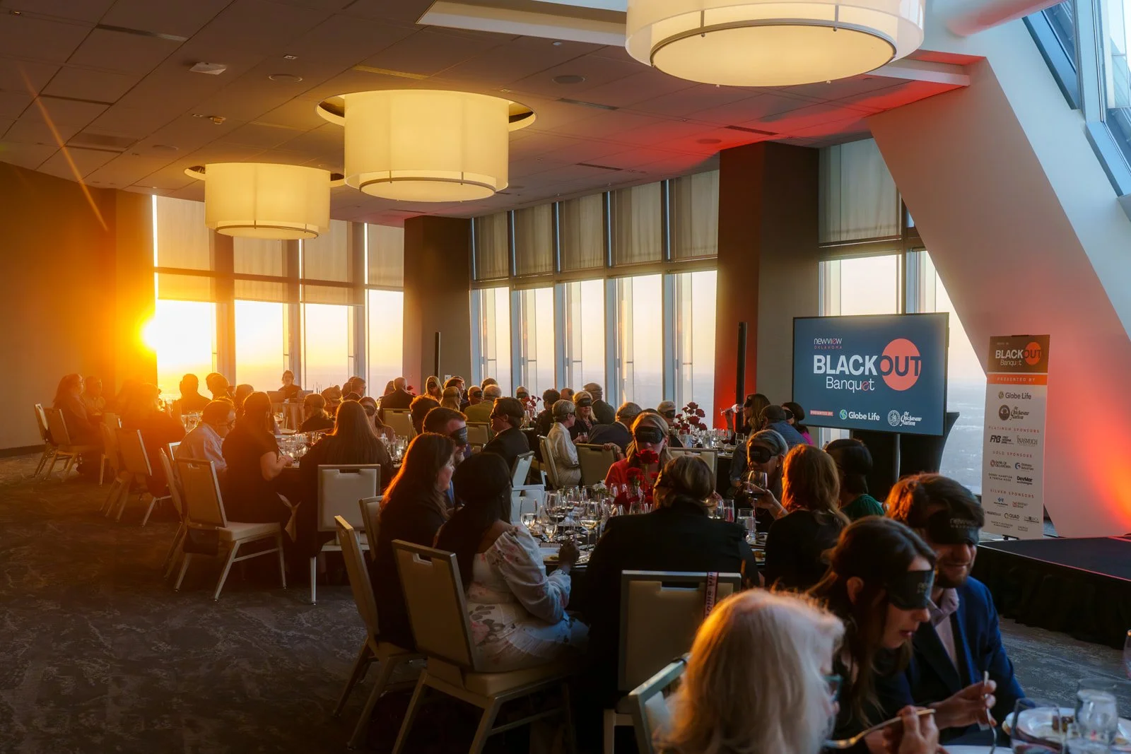 A large banquet room filled with people seated at tables during sunset, with large windows showing the sun setting over a city skyline, and a screen with 'Blackout Banquet' displayed.
