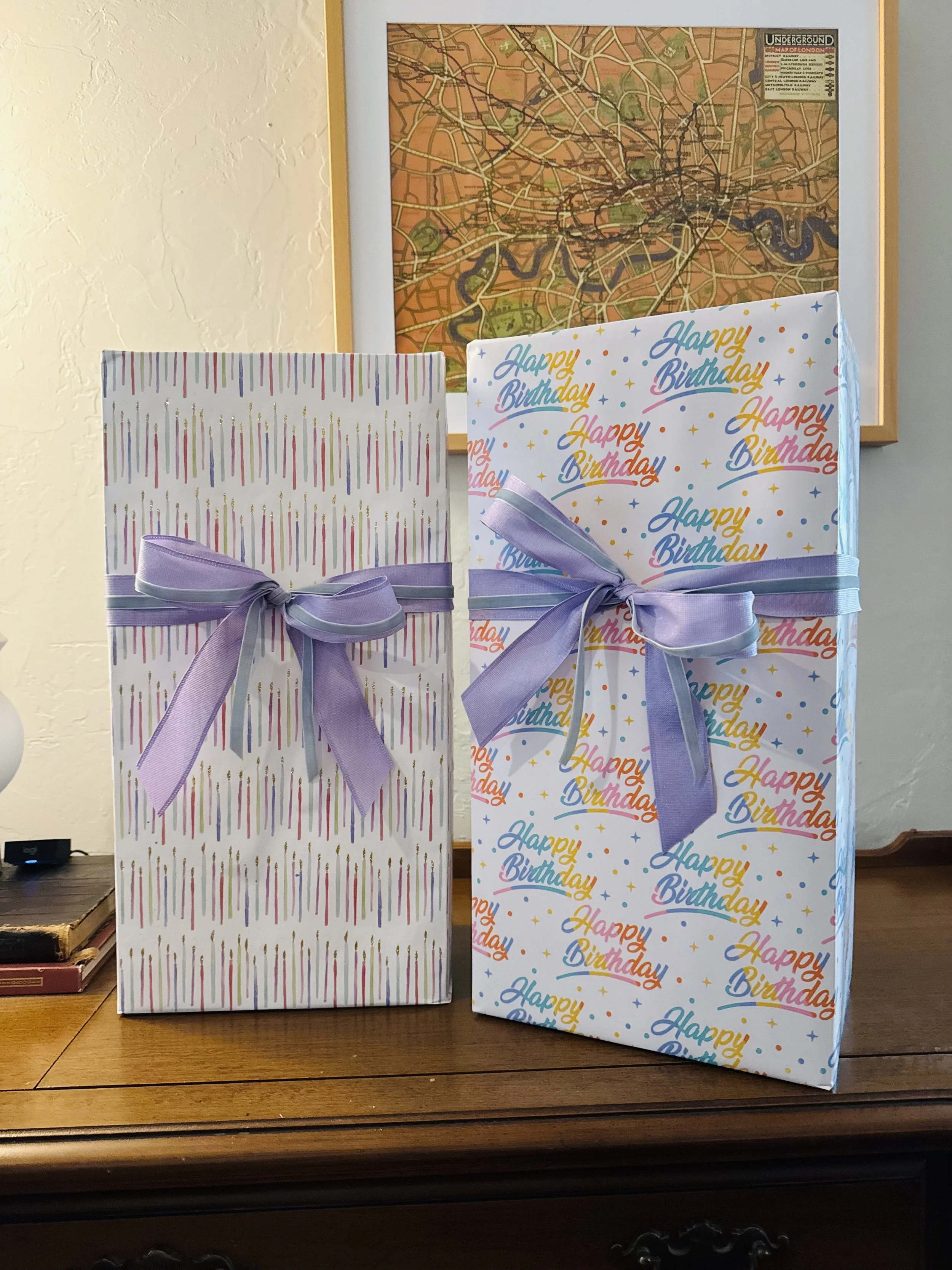 Two birthday gift bags with purple ribbons on a wooden surface, one with multicolored vertical stripes and the other with 'Happy Birthday' text in bright colors and multicolored stars, standing in front of a framed city map.