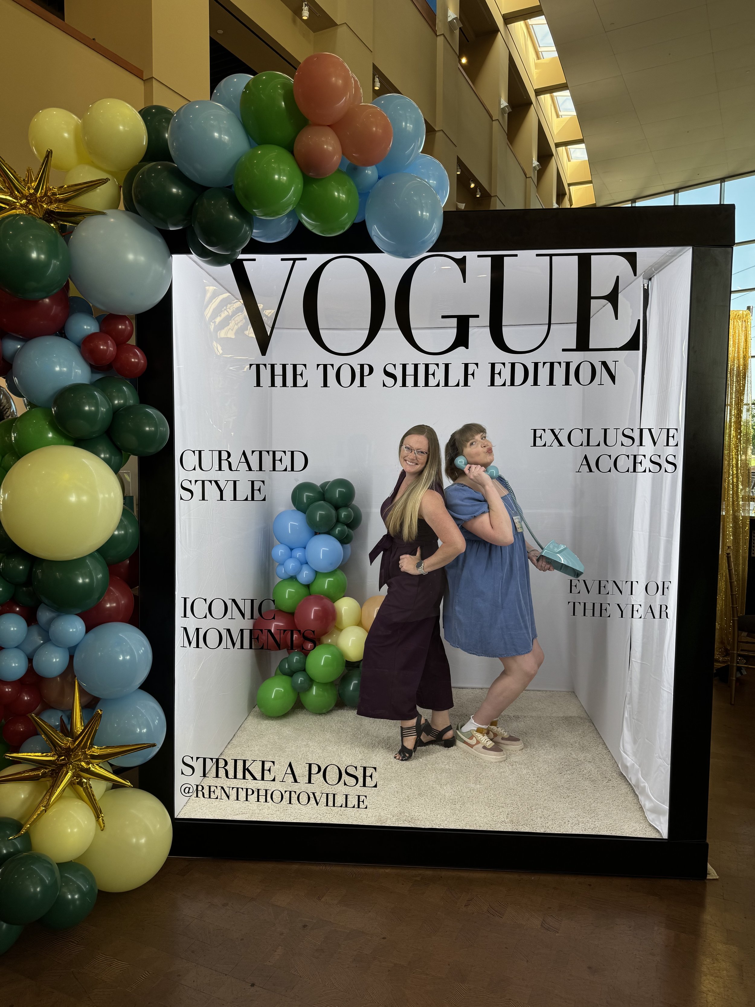 Two women posing inside a photo booth with a backdrop reading 'VOGUE THE TOP SHELF EDITION' and balloon decorations on the side. One woman is smiling and the other is holding a vintage telephone.