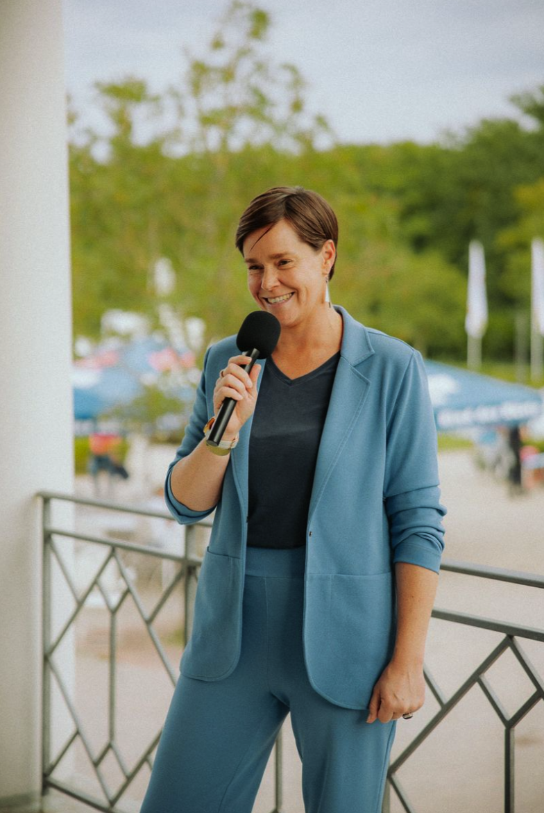 Frau in blauer Anzug hält Mikrofon und spricht im Freien, lächelnd.