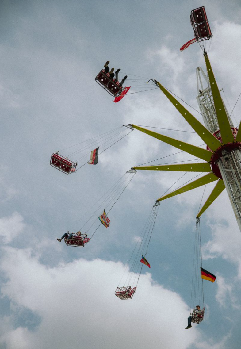 Fünf auf einem Riesenrad schaukelnde Personen, einige halten Flaggen, gegen bewölkten Himmel.