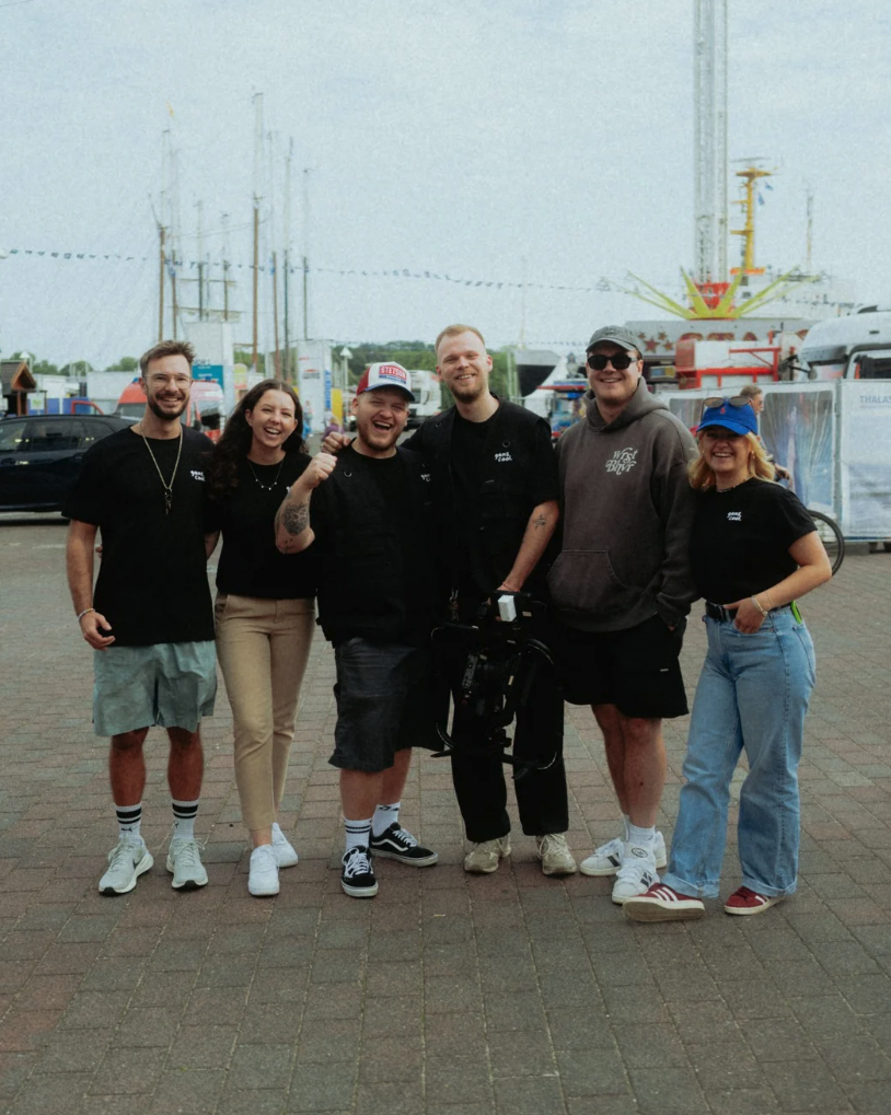 Eine Gruppe von sechs jungen Menschen steht lachend und posierend auf einem Jahrmarkt, im Hintergrund sind Fahrgeschäfte und Stände sichtbar.