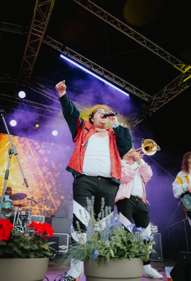 Ein Musiker singt mit einem Mikrofon auf einer Bühne, umgeben von anderen Musikern, Blumen und einer bunten Lichtshow.