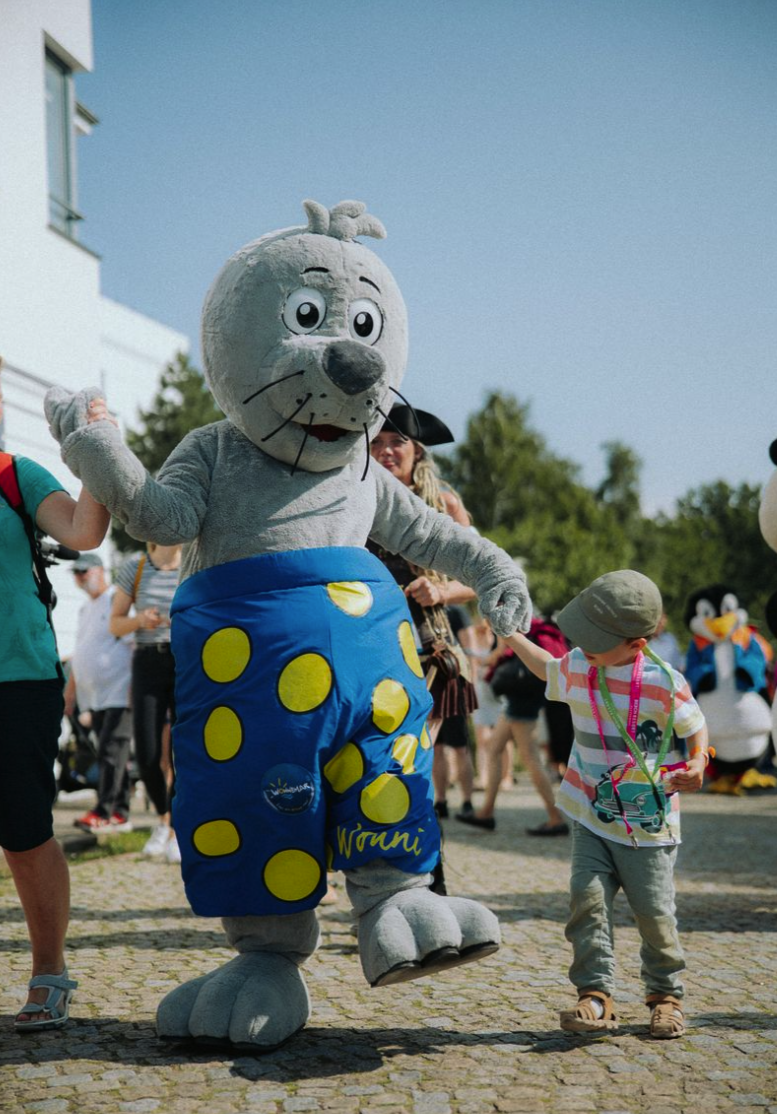 Ein Kind hält die Hand eines großen Mascots, der einen anthropomorphen Hund oder Katze in einem Sommeroutfit darstellt, bei einer Veranstaltung im Freien an einem sonnigen Tag, mit mehreren Menschen im Hintergrund.