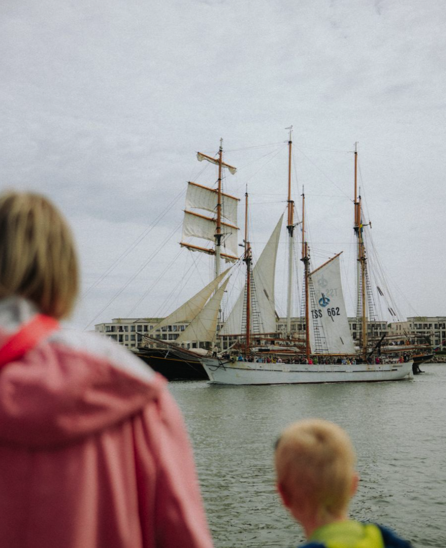 Zwei Kinder schauen auf ein Segelschiff, das im Wasser vor einem modernen Gebäude im Hintergrund fährt, bei bewölktem Himmel.