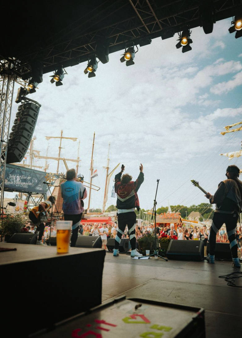 Live-Musik auf einer Bühne bei einem Open-Air-Konzert mit Musikern, die vor Publikum spielen, während die Sonne scheint und Wolken am Himmel sind.