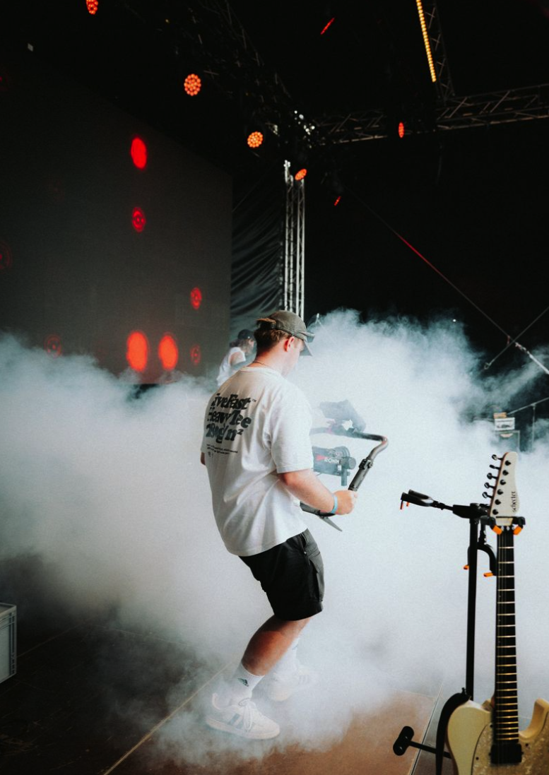 Musiker auf einer Bühne vor einem großen Bildschirm, umgeben von Nebel und rotem Licht, mit einer Gitarre im Hintergrund.