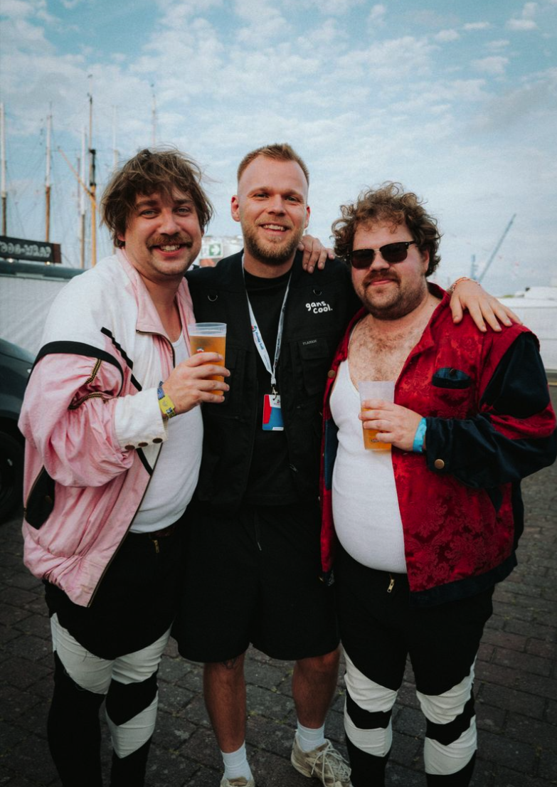 Drei Männer stehen nebeneinander, lachen und halten Bier. Der Hintergrund zeigt ein Hafen mit Segelschiffen.