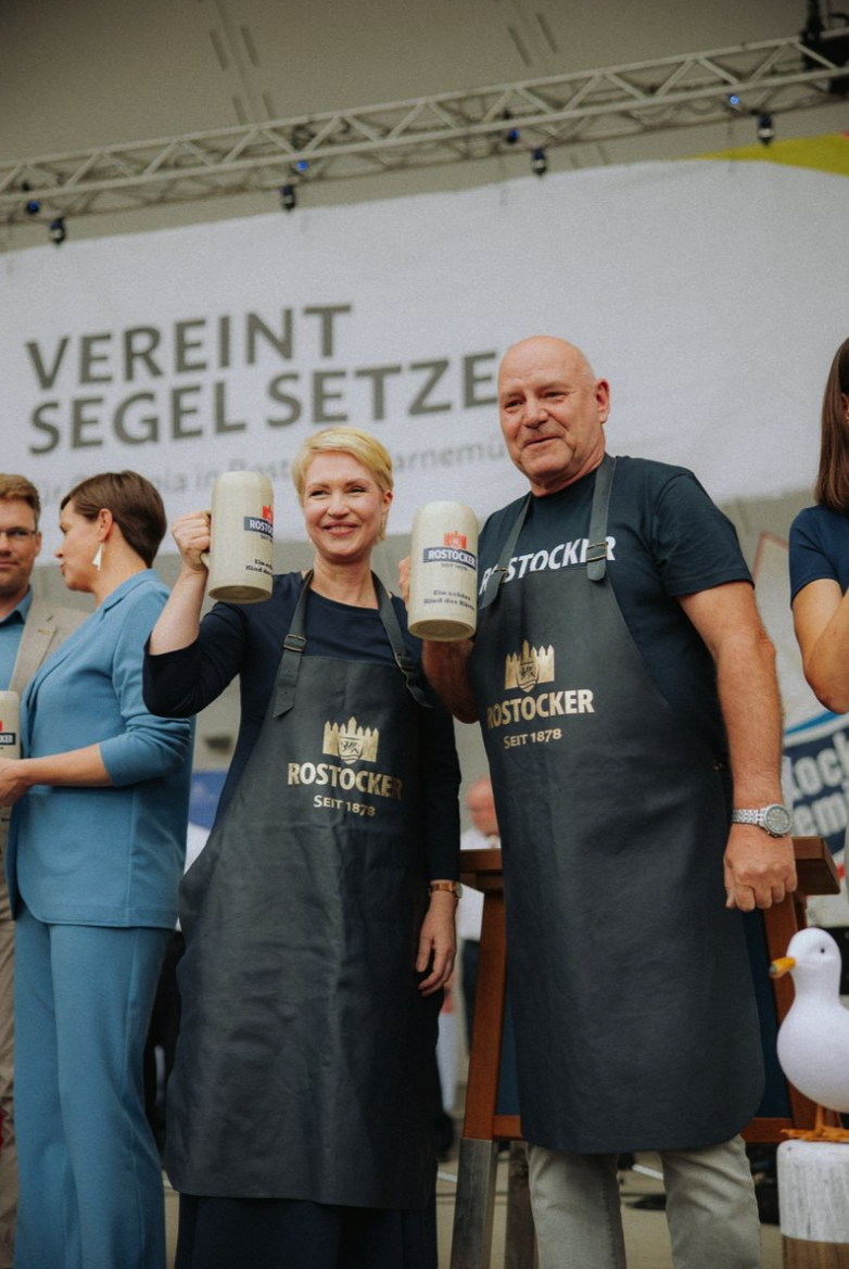 Menschen bei einem Event, die Bierkrüge hochhalten, mit einem Hintergrundtext "VEREINT SEGEL SETZE".