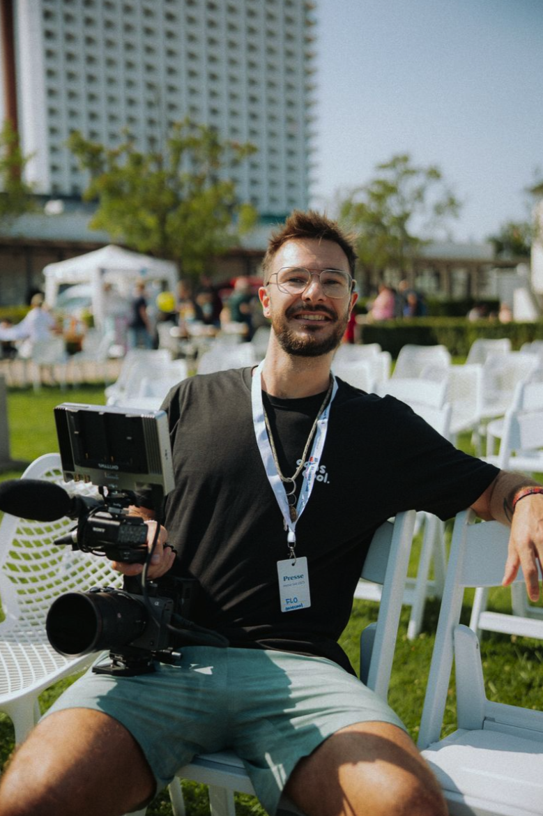 Ein junger Mann mit Brille und Bart sitzt im Freien während eines Events, hält eine Videokamera in der Hand und trägt einen Ausweis mit der Aufschrift 'Presse'.