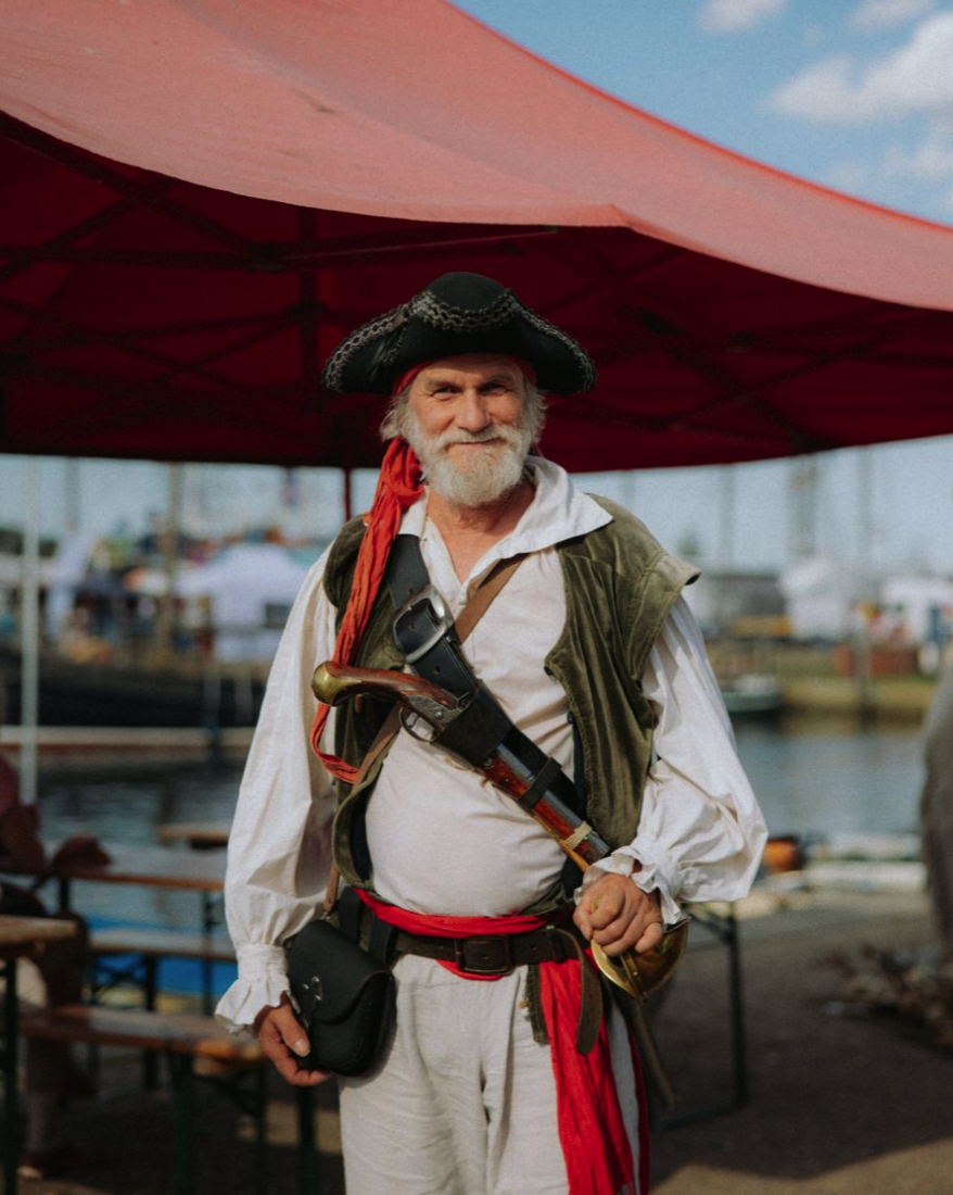 Ein älterer Mann in Piratenkostüm mit Hut, Beil und einer Pistole, die an seinem Gürtel hängt, steht am Hafen unter einem roten Sonnensegel.