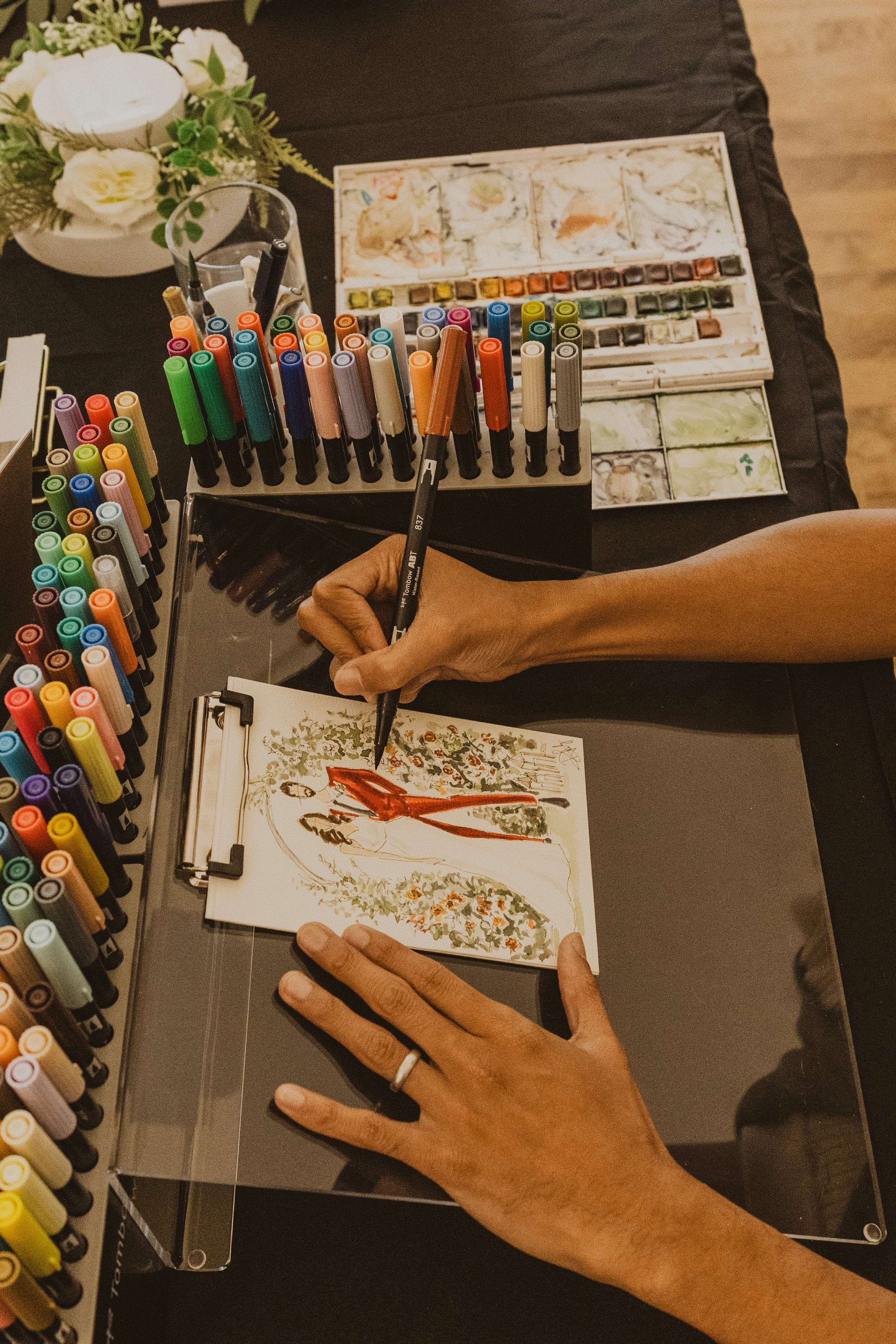 Person creating watercolor artwork of a woman in a red dress with floral decorations around her on a sketchpad on a black table, surrounded by colorful markers and watercolor paints.