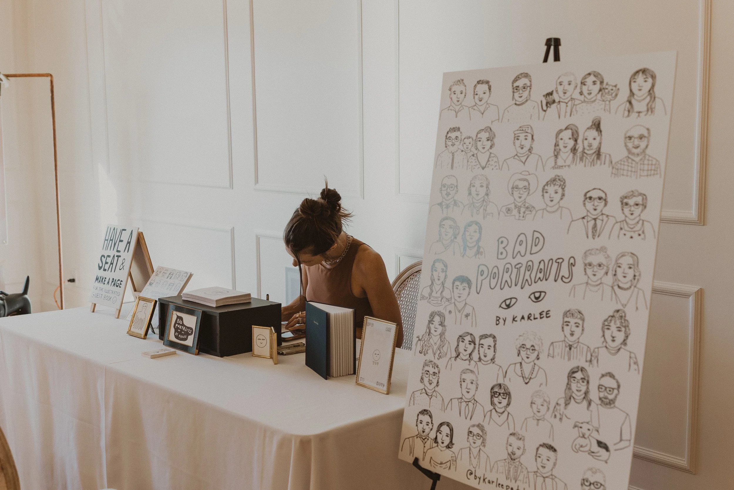 A woman at a table with books and framed pictures, sitting beside a large poster showcasing illustrated portraits of various people, titled 'Bad Portraits by Karlee'.