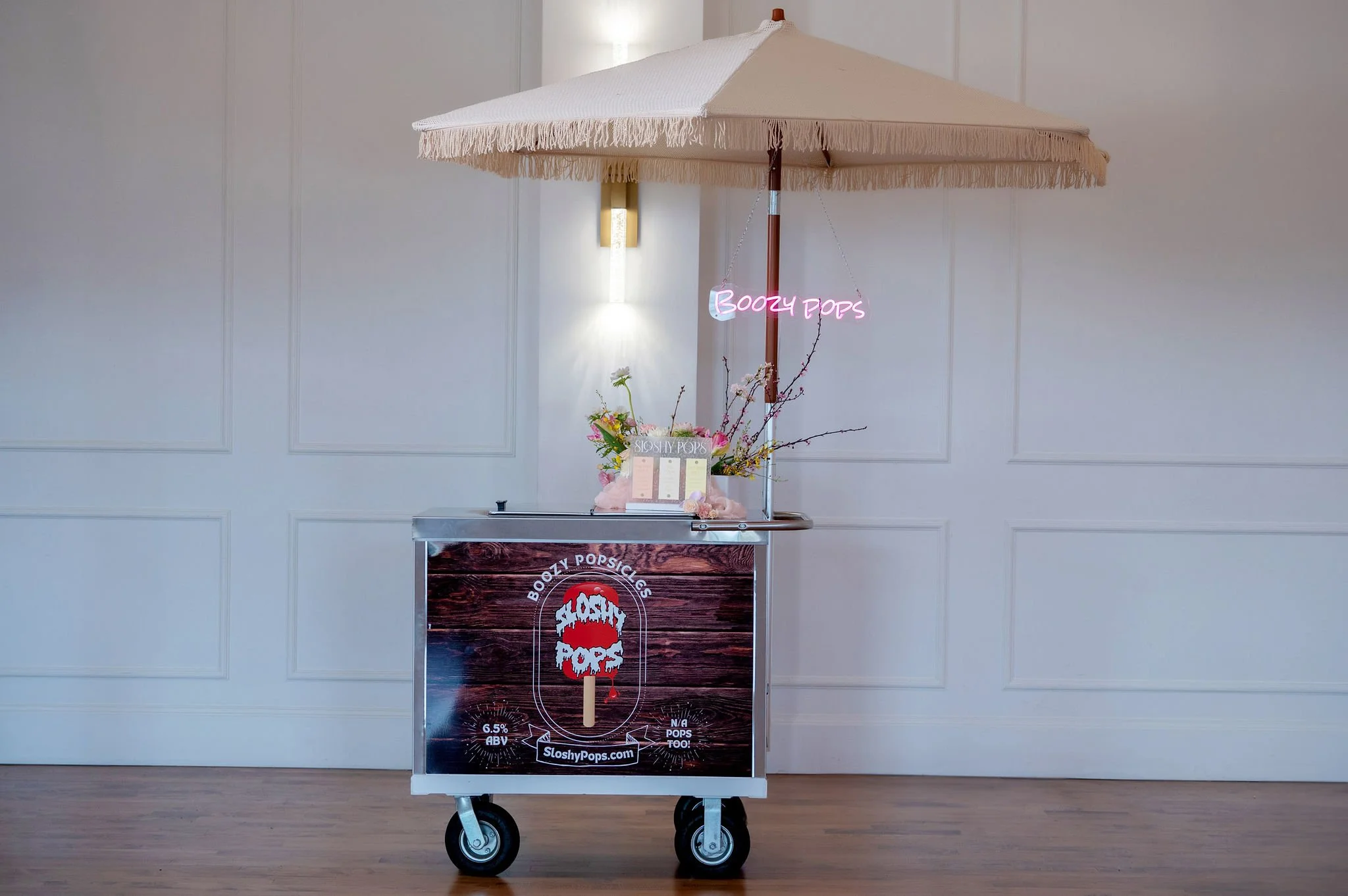 A popsicle cart with a purple neon sign that reads "Boozy Pops" and a floral decoration on top. The cart is on wheels and has a black table with a menu. The background features a white wall with decorative paneling.