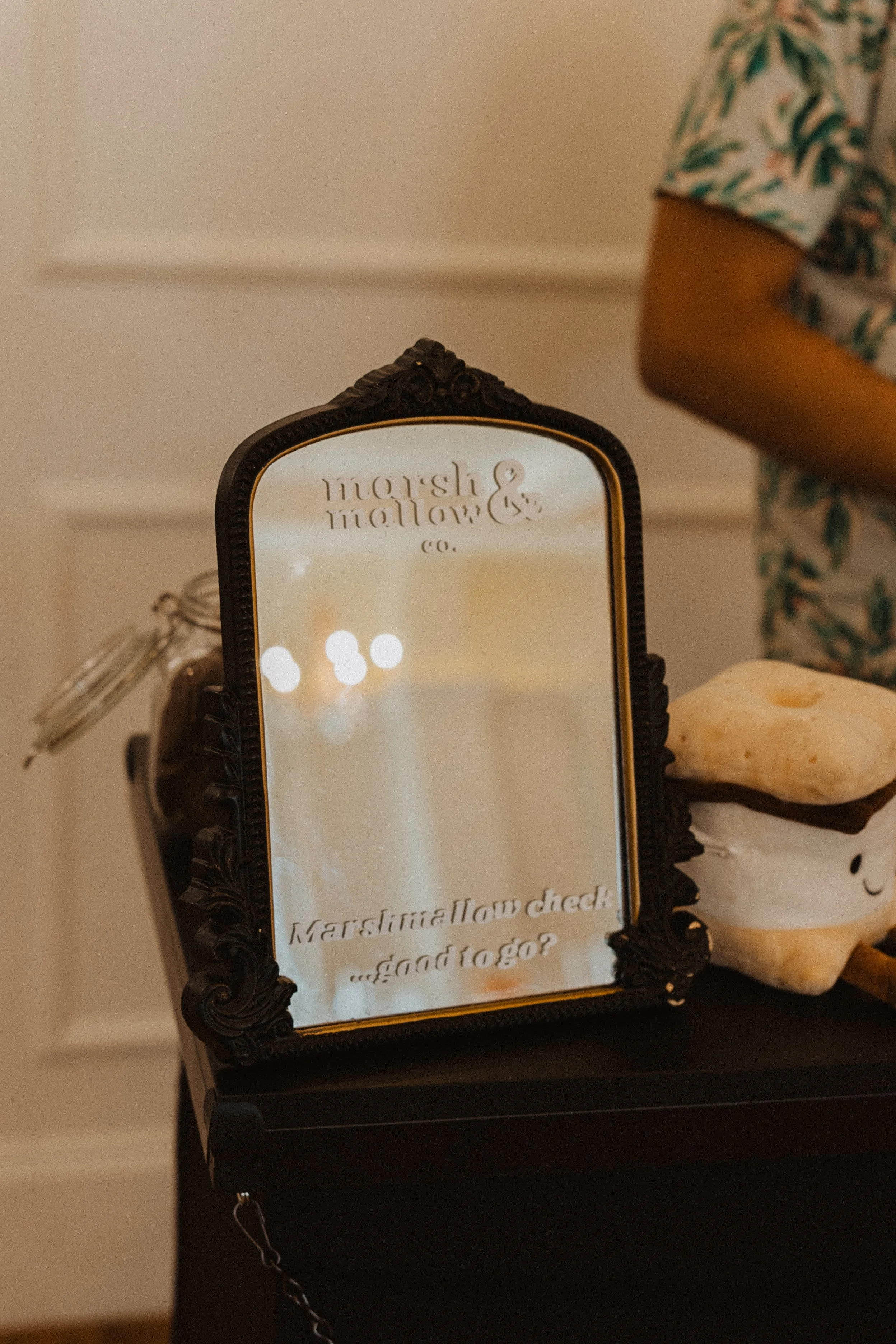 A vintage-style black frame mirror with the words "marsh &mallow & co." engraved at the top, placed on a black surface with a nearby white skull-shaped object and a rock, and a person wearing a short sleeve shirt with a colorful leaf pattern in the background.