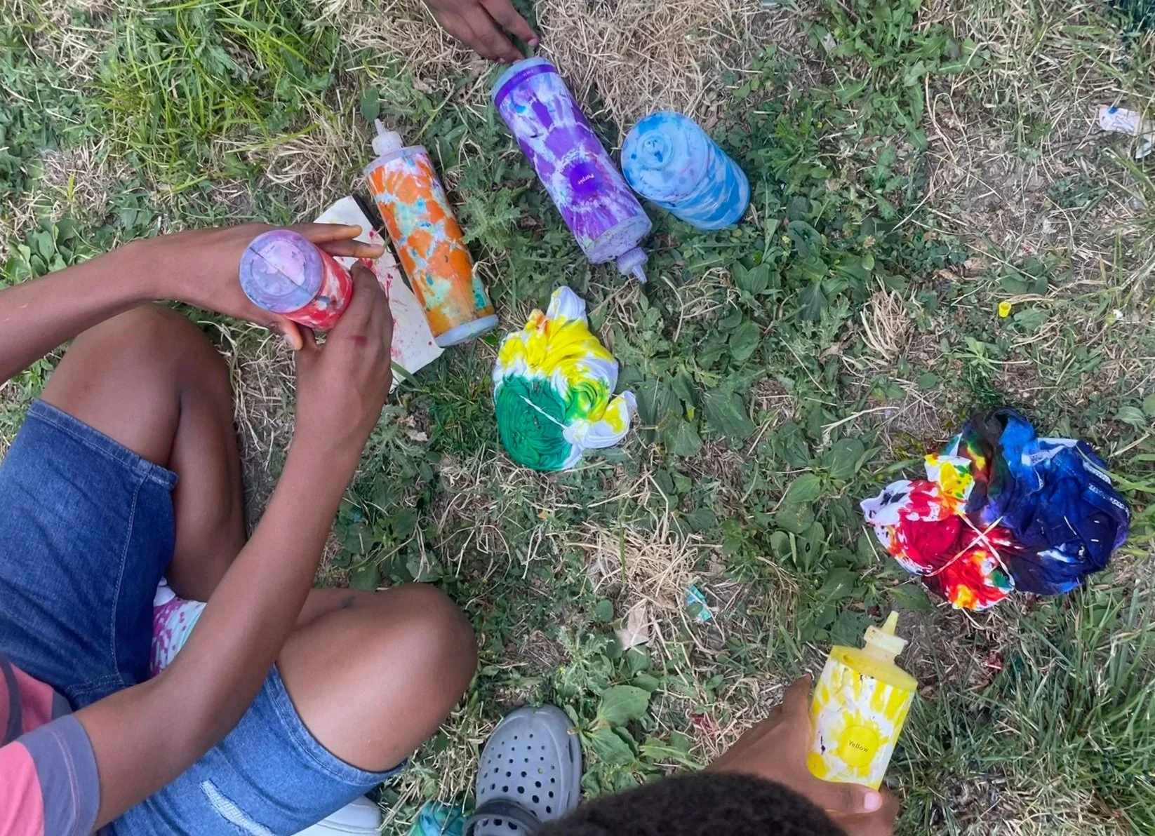 Children outdoors on grass painting with colorful water-based paints, with paint bottles, cups, and tissue paper scattered around.