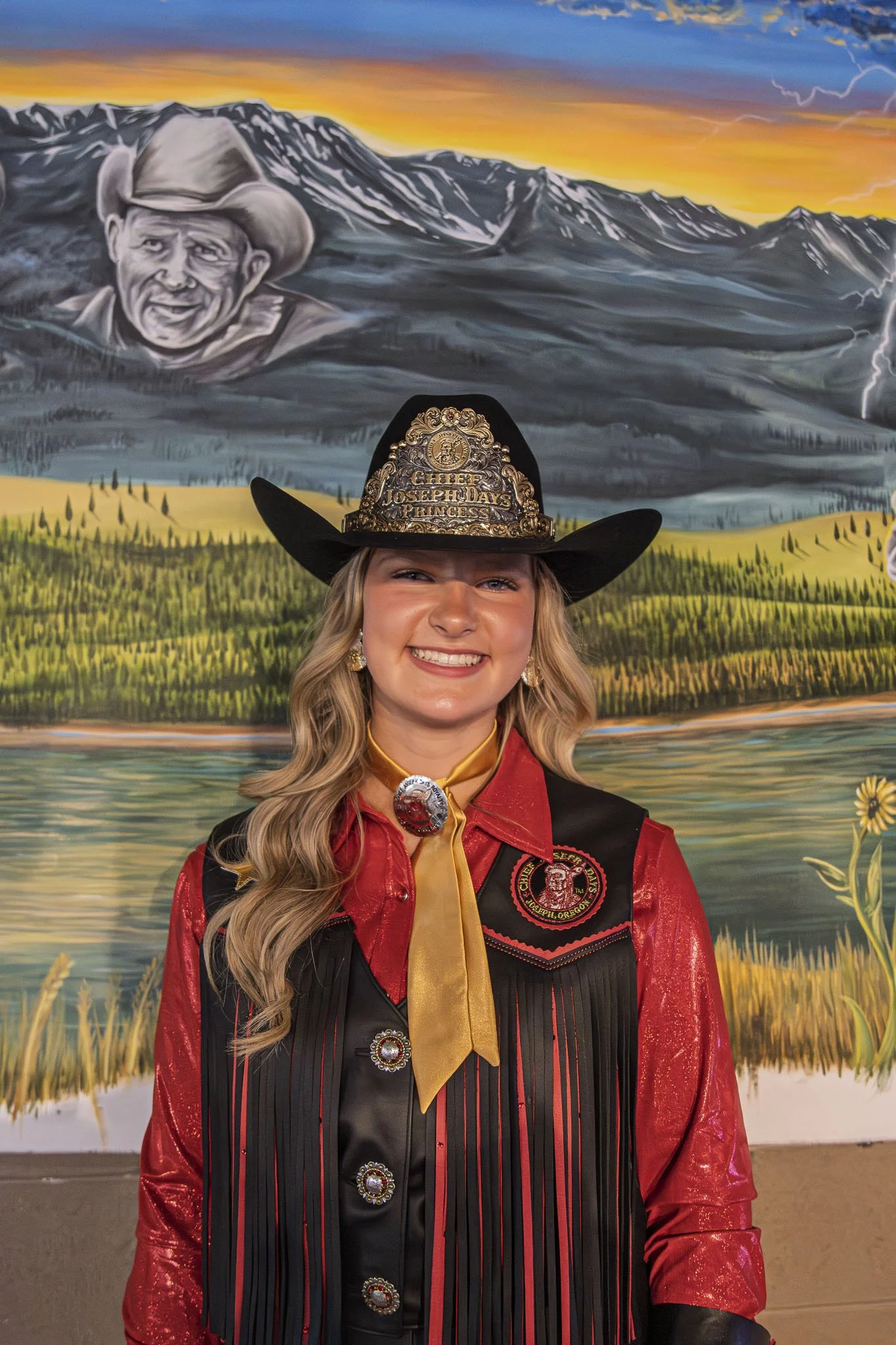 Cowgirl wearing a western hat with fringe vest and red shirt, smiling against a wooden background.