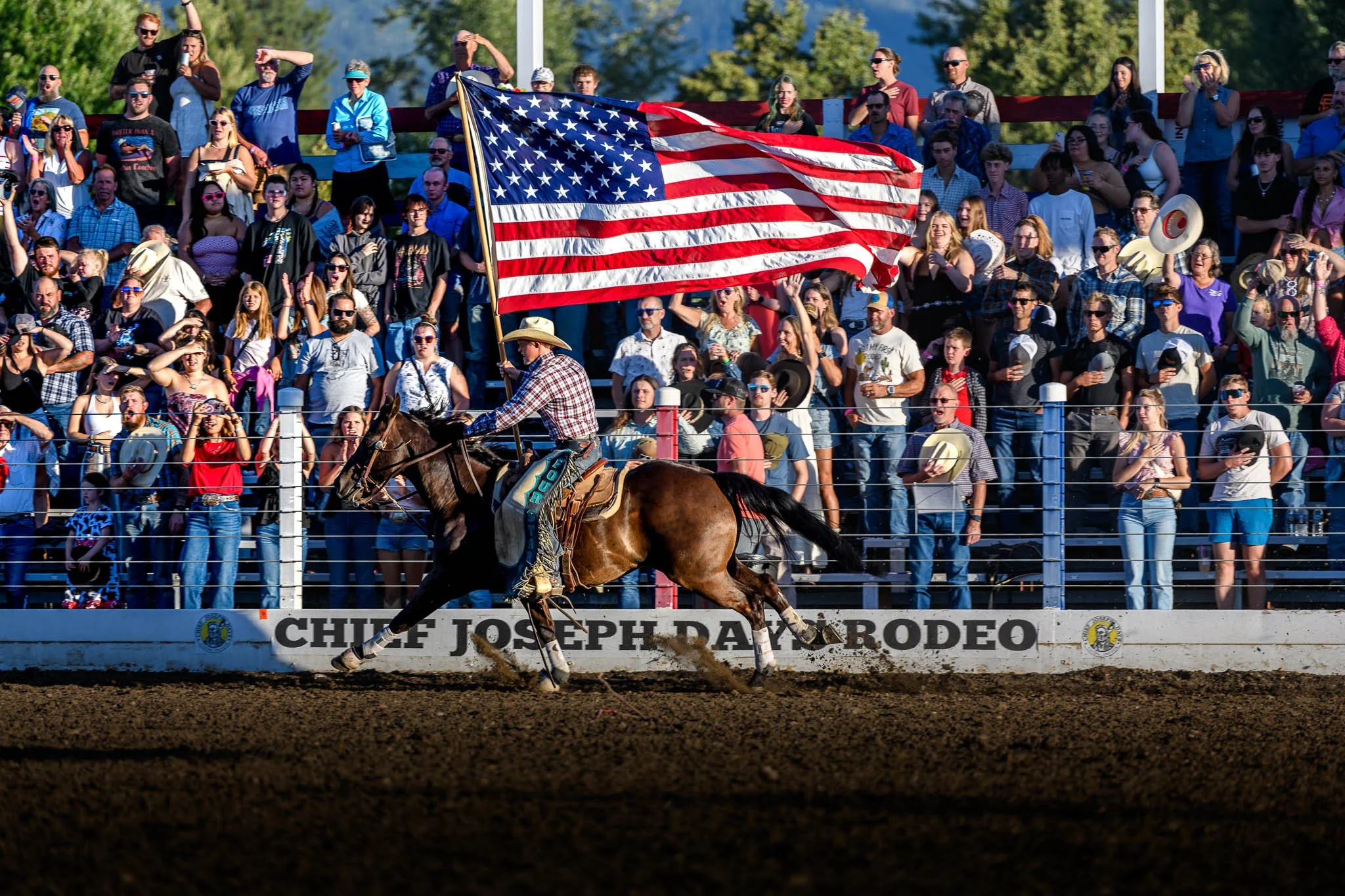 A cowboy riding a horse at Chuck Joseph Day Rodeo, carrying a large American flag, with a crowd of spectators in the background.