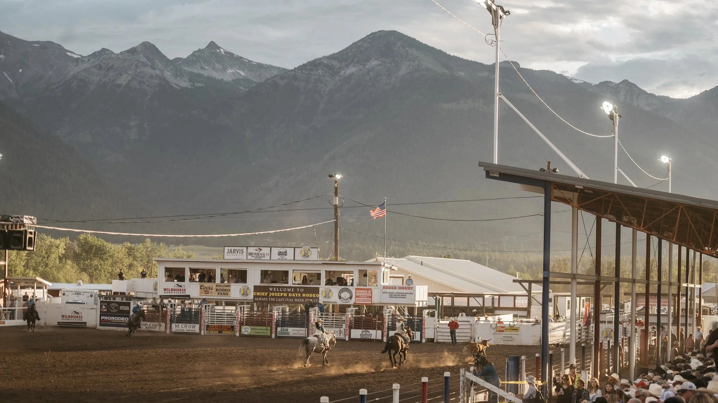 Rodeo arena with cowboys on horseback, spectators watching, and mountains in the background.