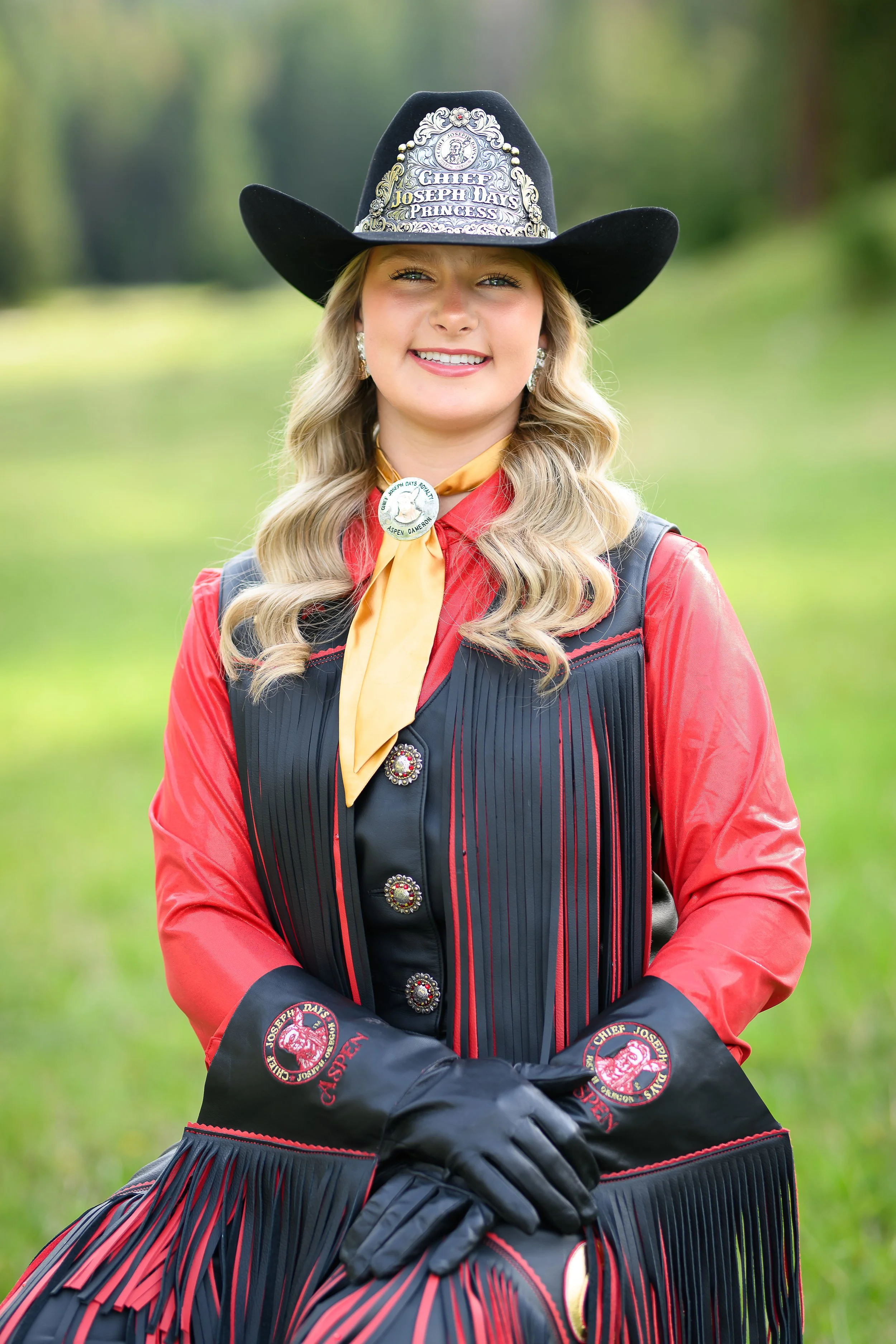 Cowgirl wearing a western hat with fringe vest and red shirt, smiling against a wooden background.