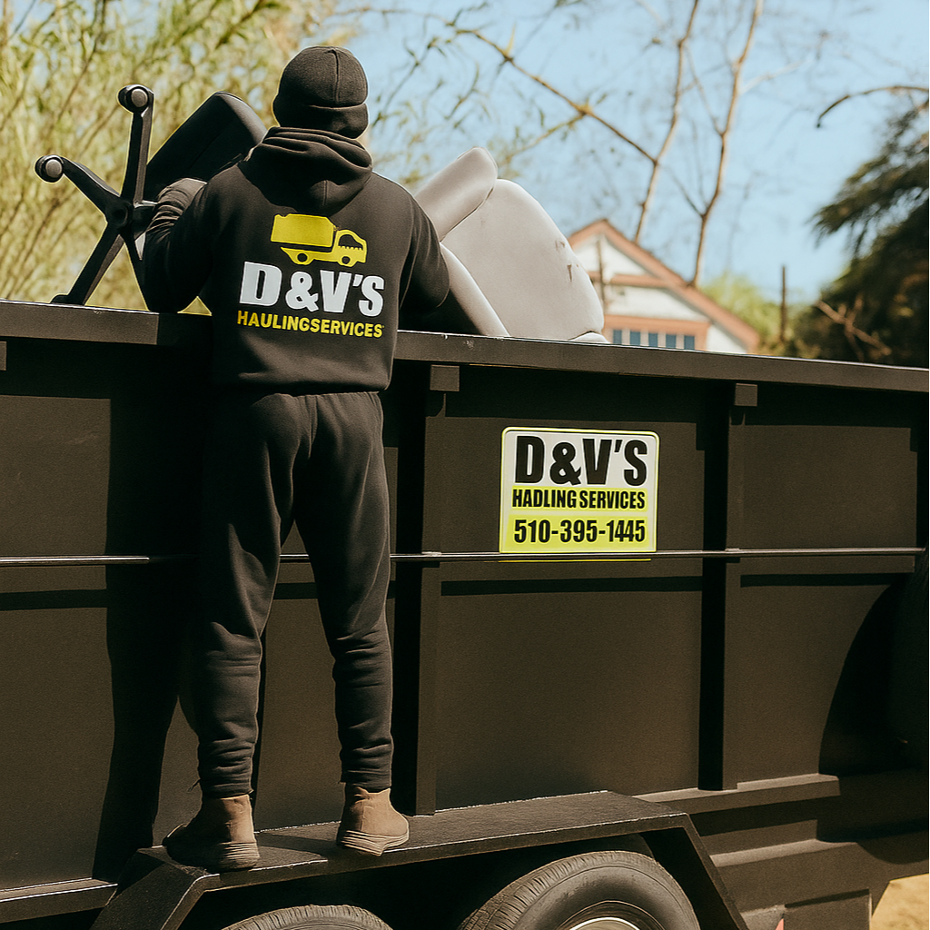 Junk removal employees stand on side of trailer and load unwanted junk into black dump trailer with logo of D&V's Hauling service on side of dump trailer. Junk removal BayPoint