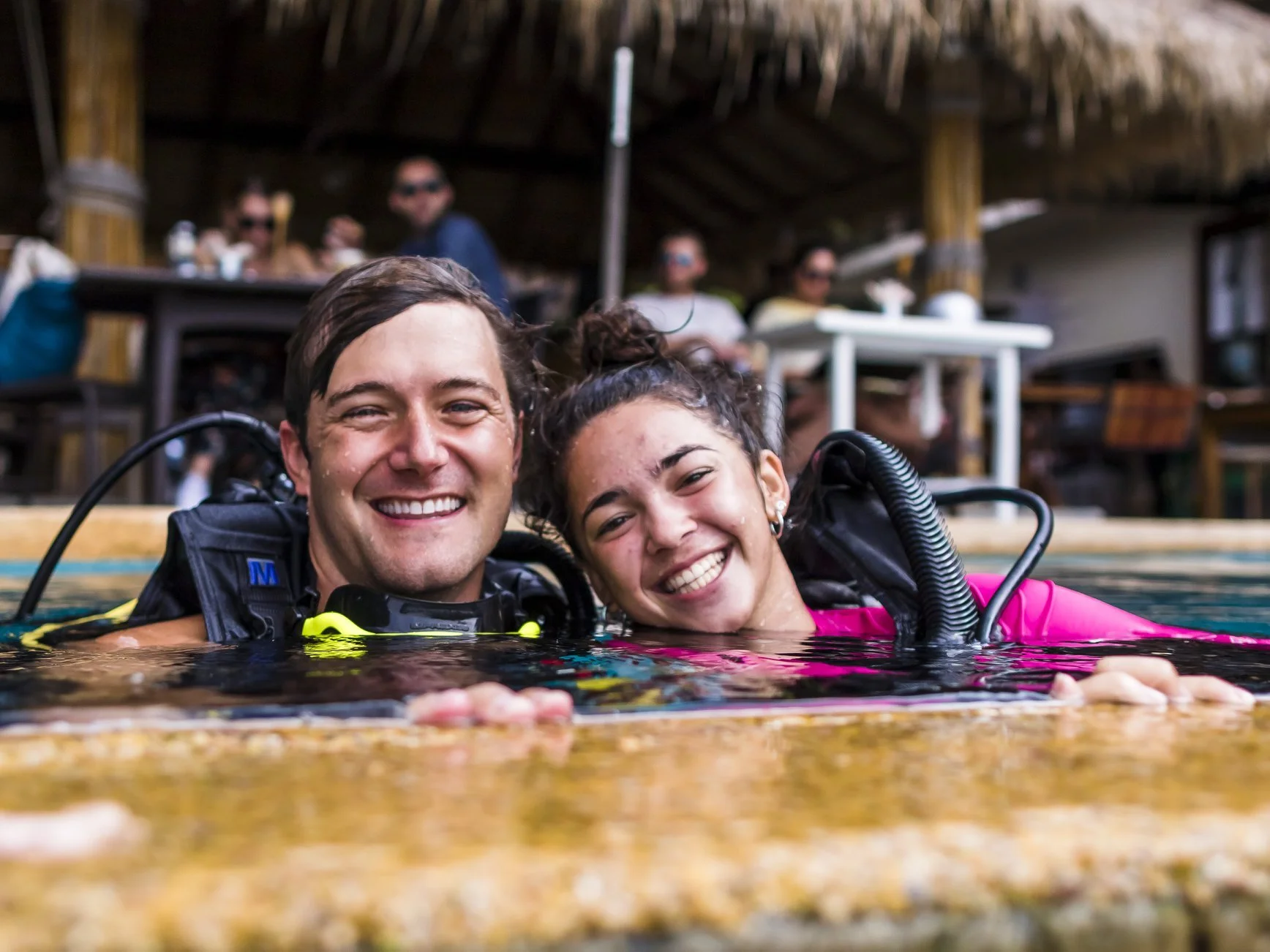 Two smiling scuba divers, a man and a woman, are partially submerged in a pool, wearing wetsuits and diving gear. They are close together, holding onto the pool edge, with a thatched-roof structure and other people blurred in the background.