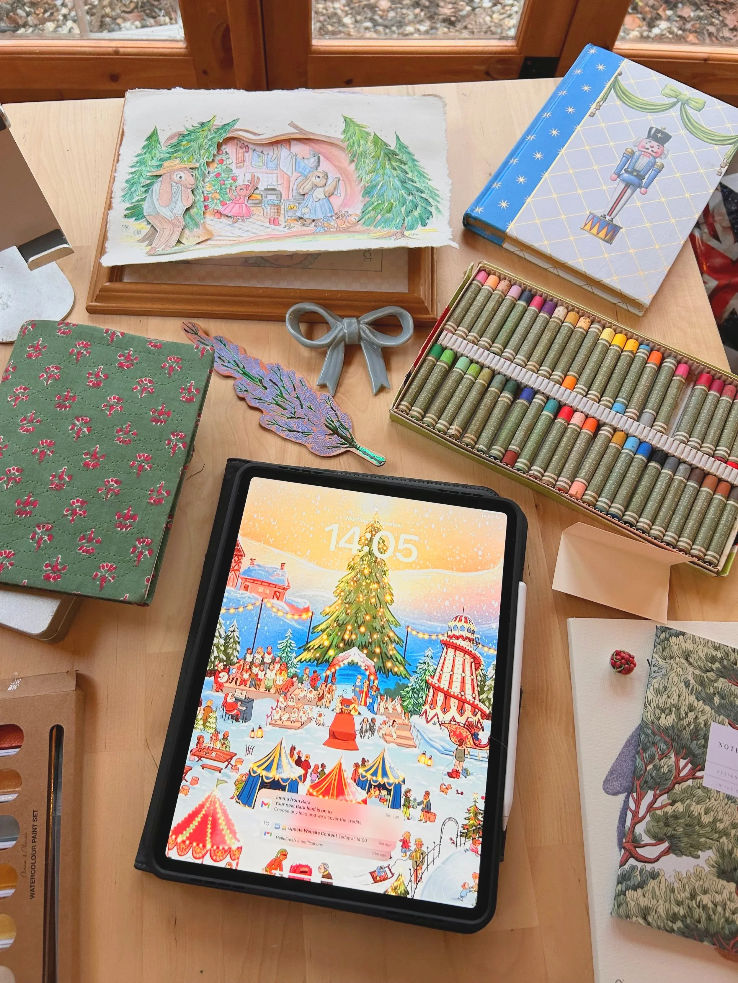 A festive illustrated holiday scene on a tablet screen showing a Christmas tree, carolers, and a carnival, with various colorful objects and holiday-themed decorations around it on a wooden table.