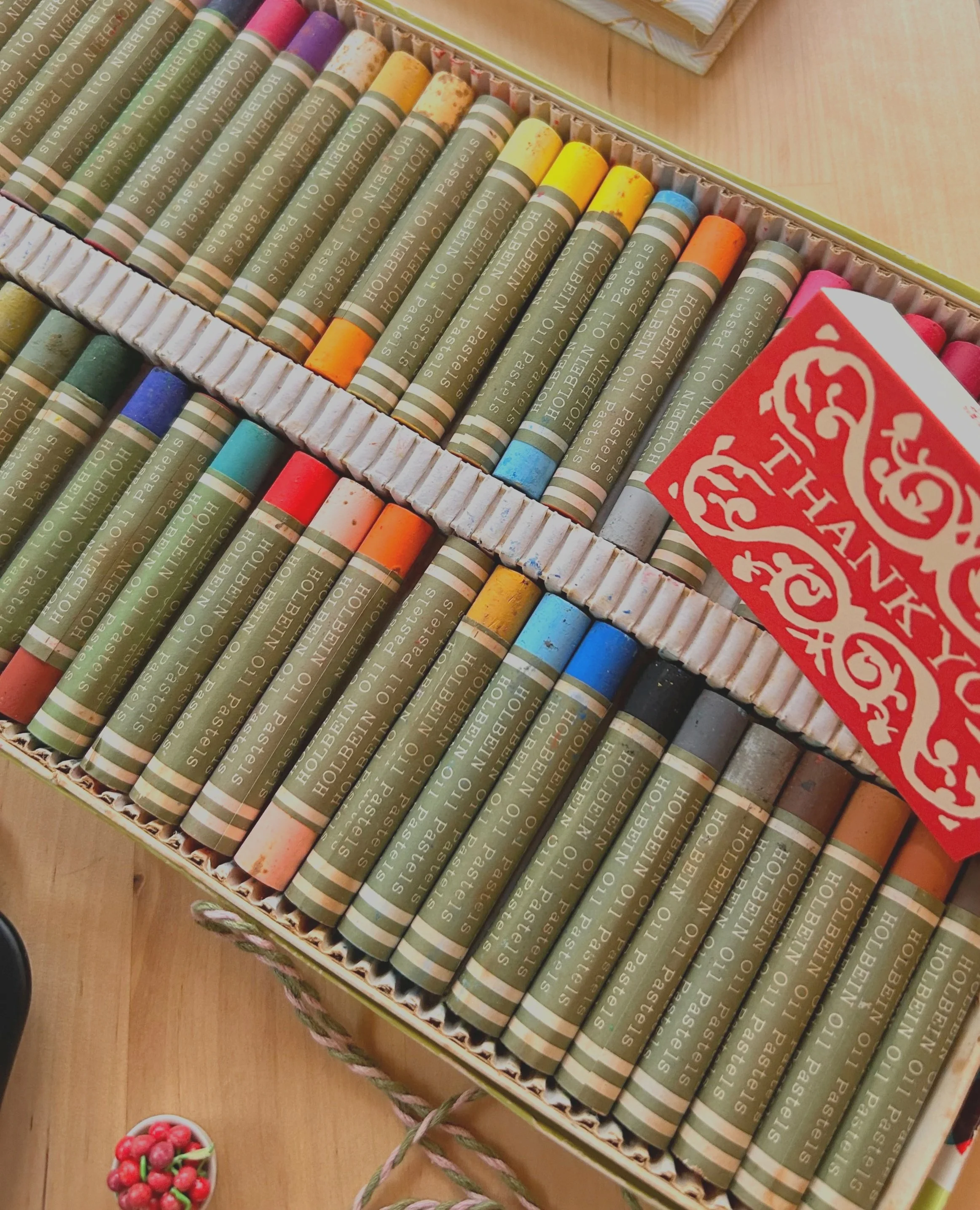 A box of colored oil pastels arranged neatly in rows on a wooden surface, with a small bowl of red berries nearby.