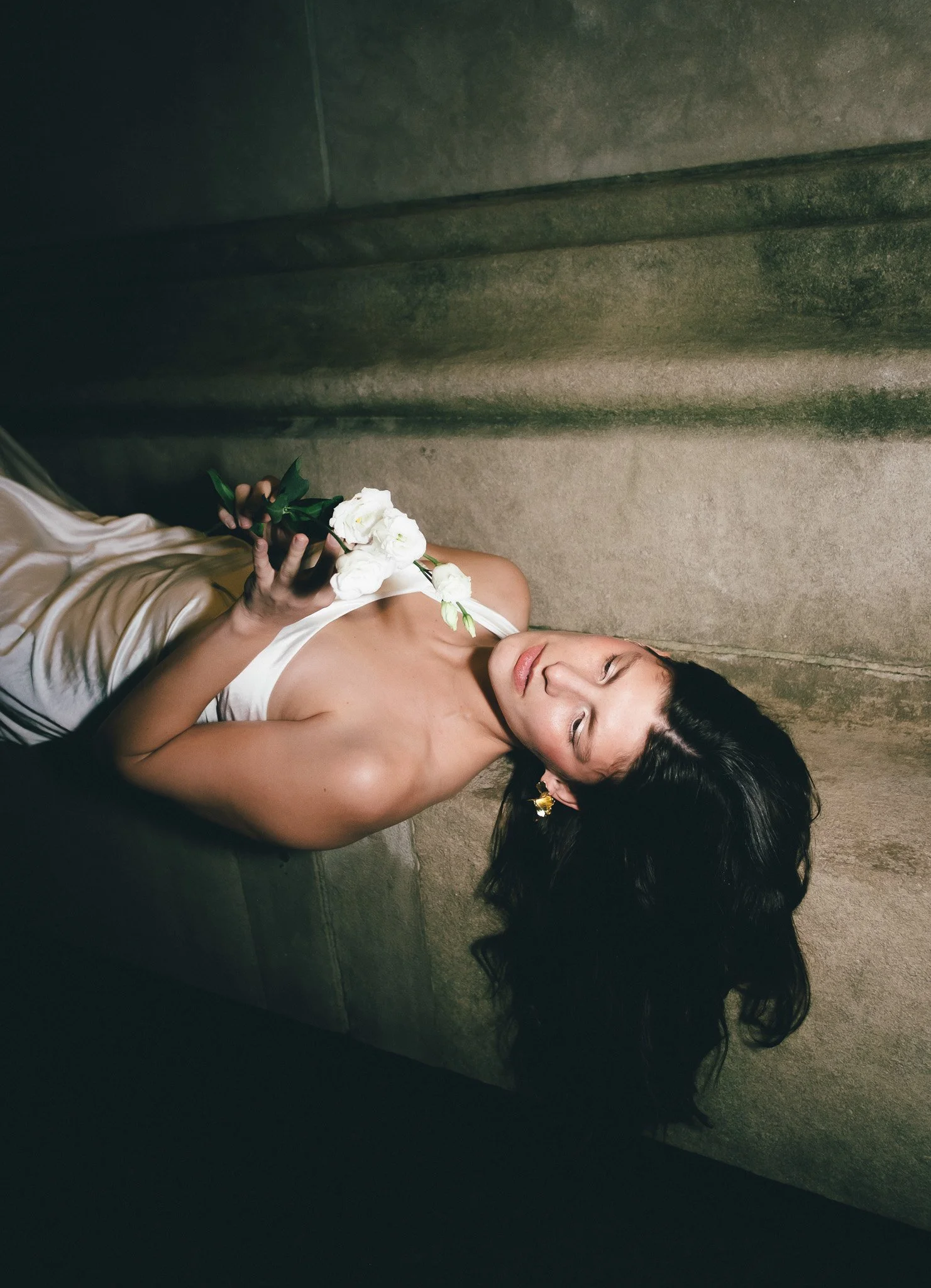 A woman lying on the floor holding a bouquet of white flowers, with her eyes closed, wearing a white satin dress and gold earrings.