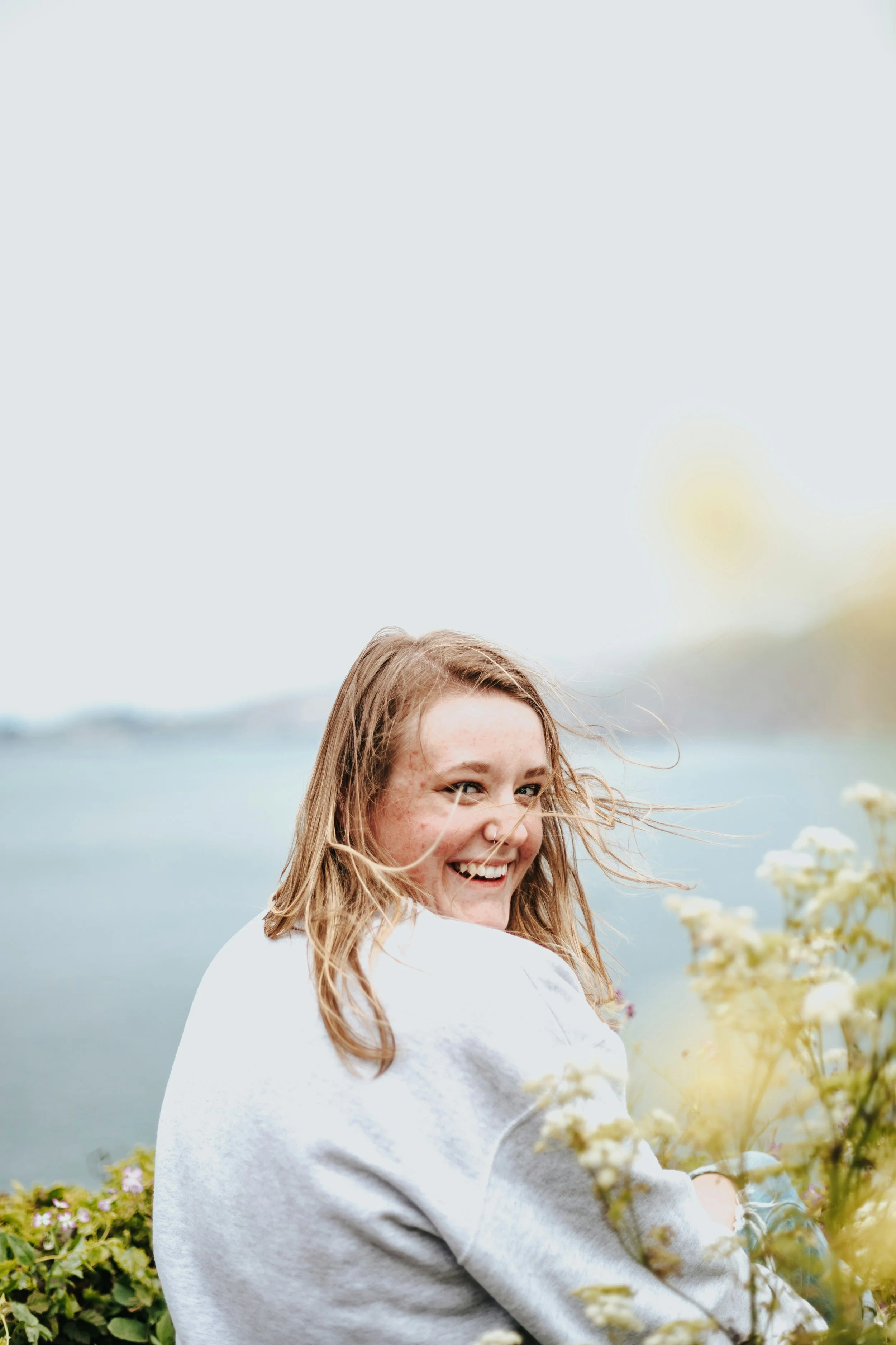 A young woman with blonde hair smiling and looking back, outdoors by a body of water with cloudy sky, surrounded by flowers.