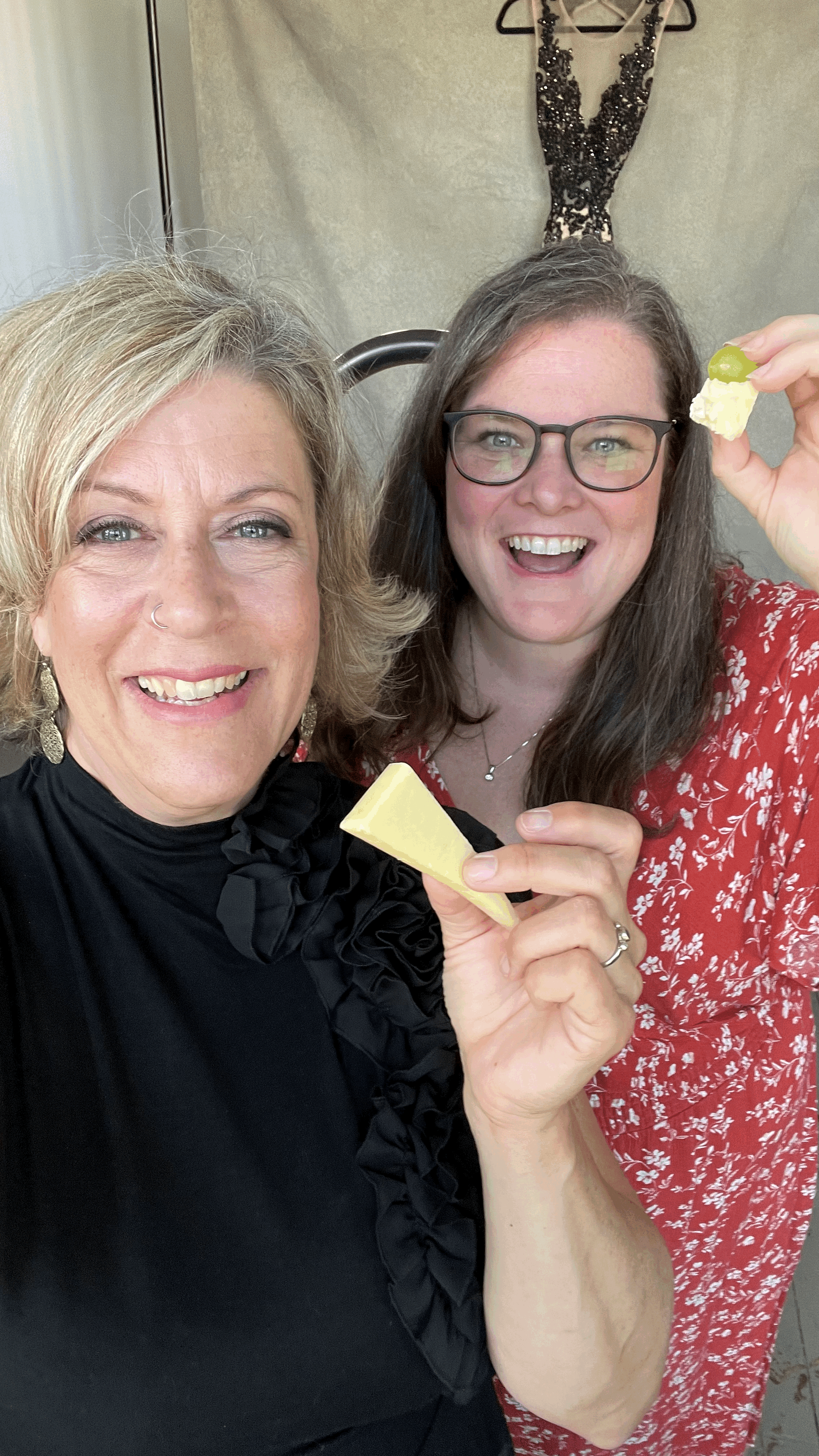 Two smiling women holding cheese and a grape in a room with beige walls and a black dress hanging in the background.