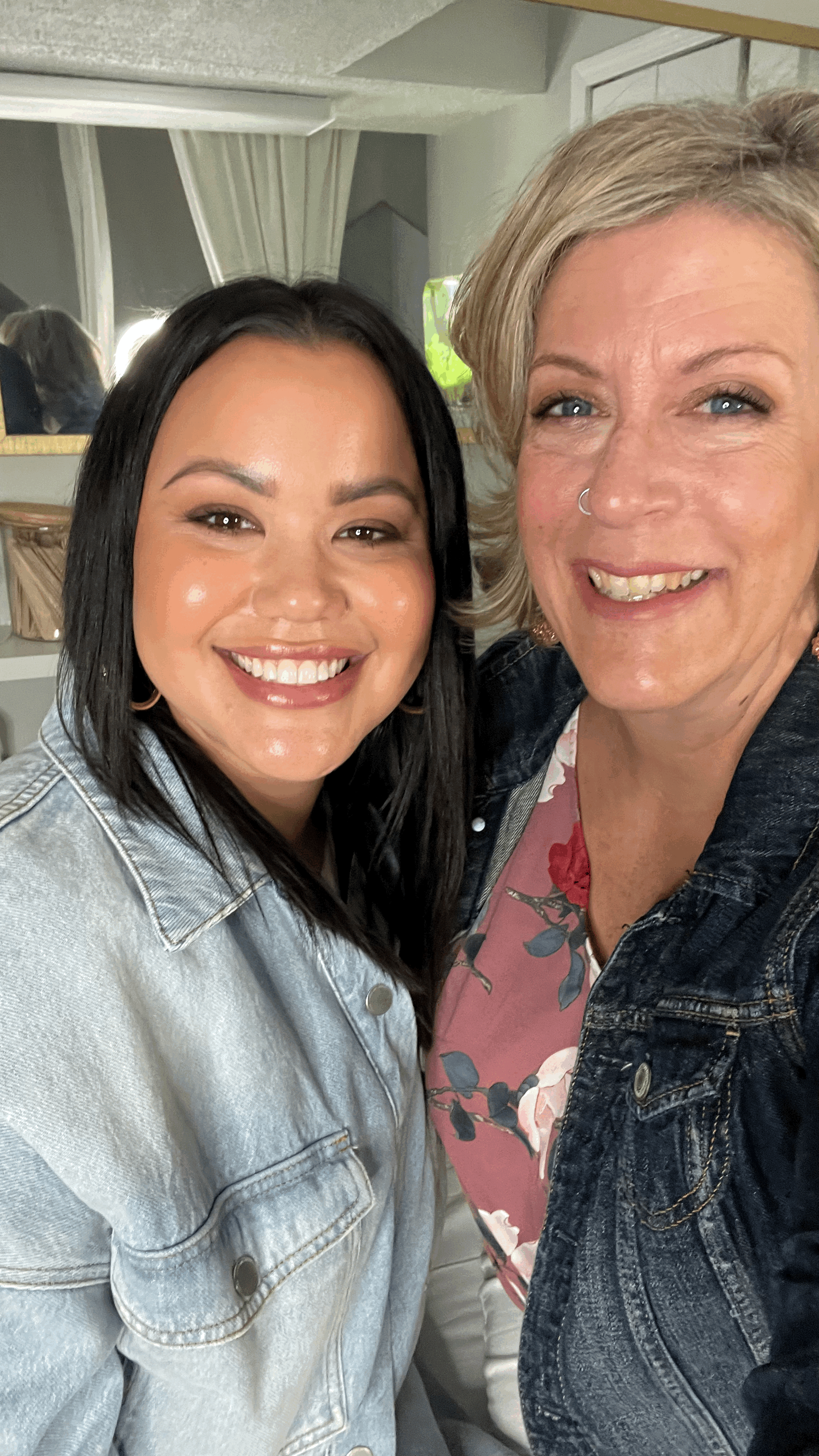 Two women taking a selfie indoors, smiling, with a mirror and curtains in the background.