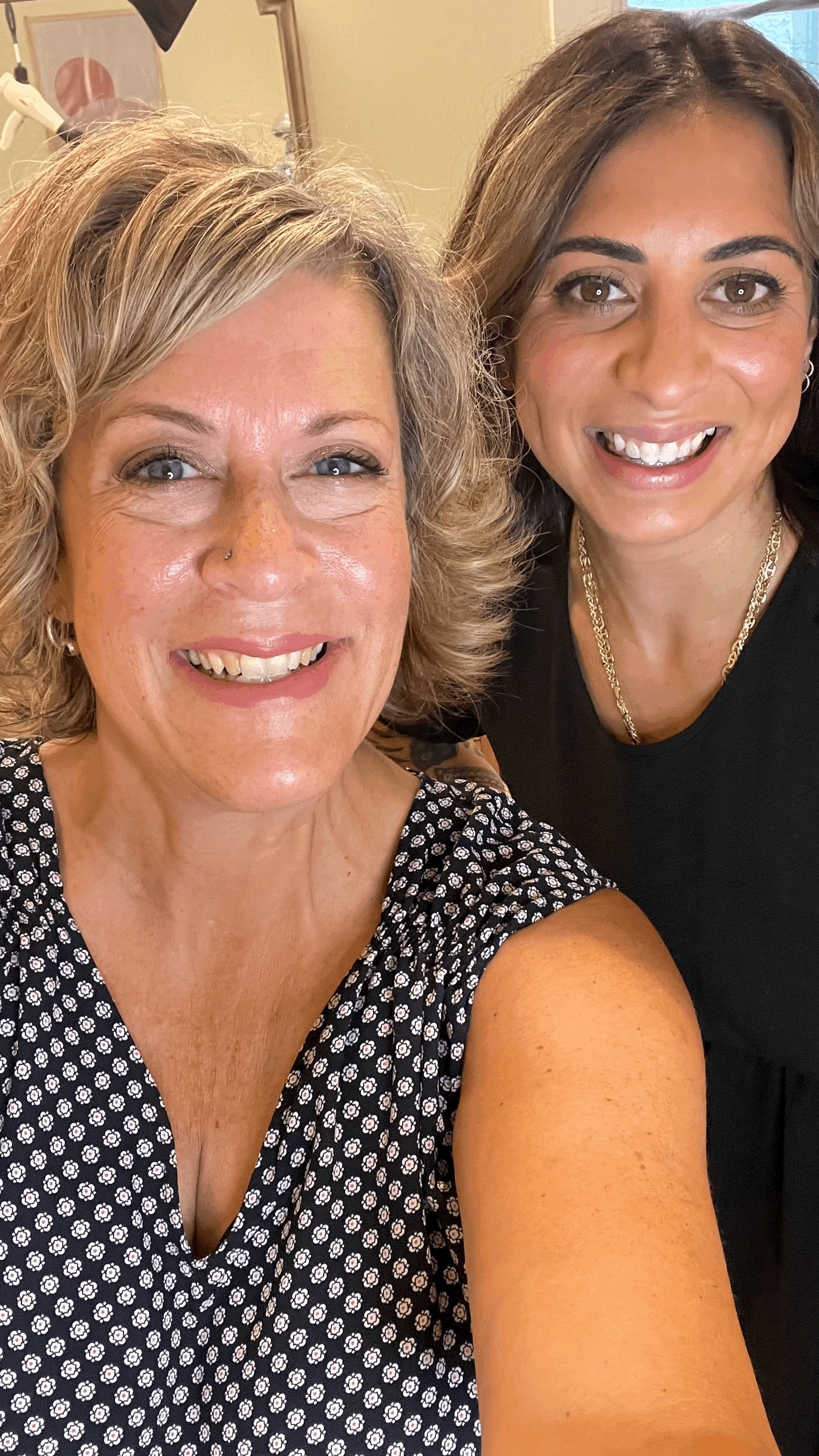 Two women smiling, one with curly gray hair and the other with straight brown hair, taking a selfie together indoors.