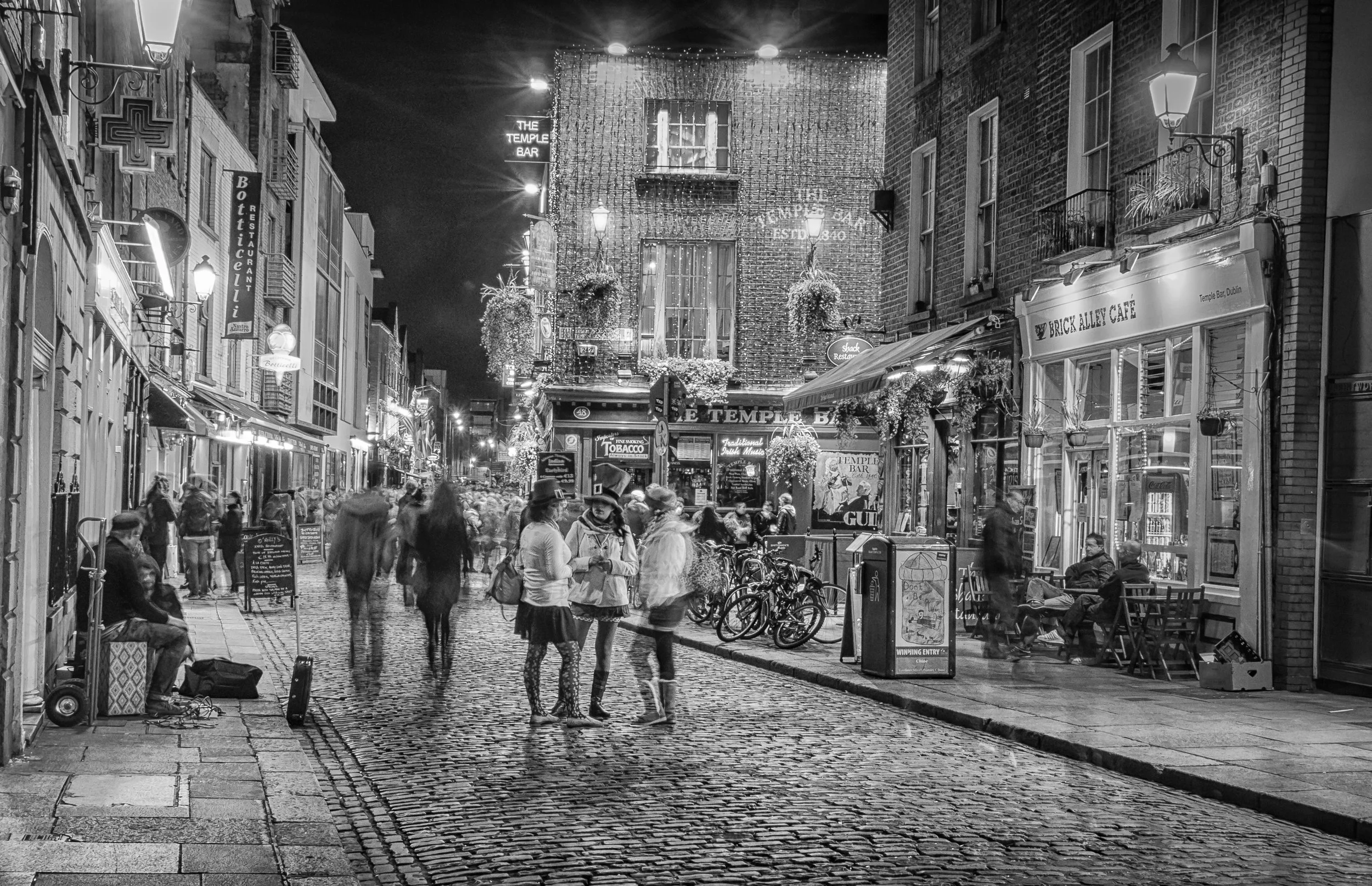 Nighttime street scene with people walking on cobblestone street, surrounded by buildings with lit signs for bars and restaurants, some outdoor seating and bicycles parked along the sidewalk.