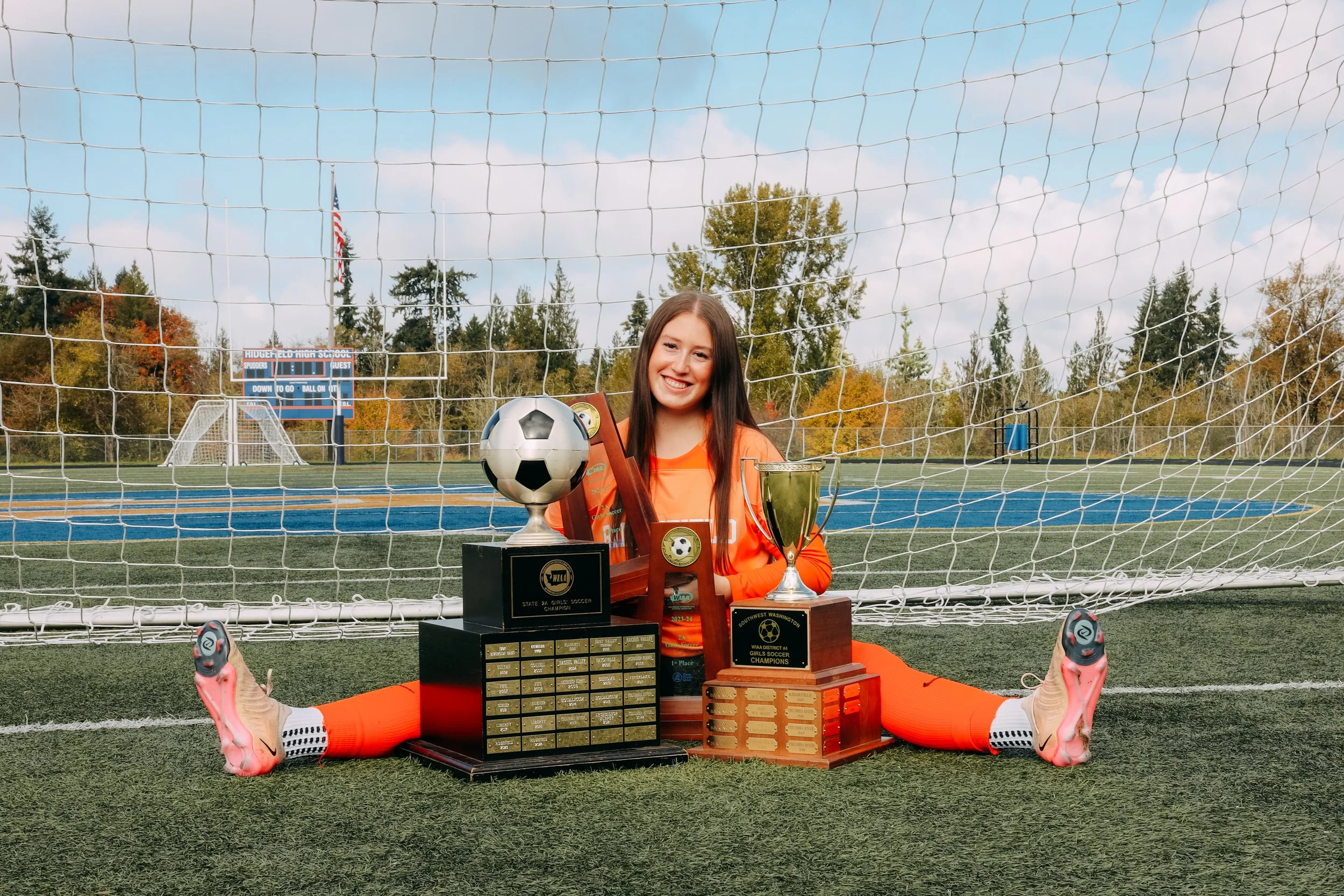 Smiling female soccer player in orange uniform sitting behind trophies and awards on a soccer field with goal net in the background.