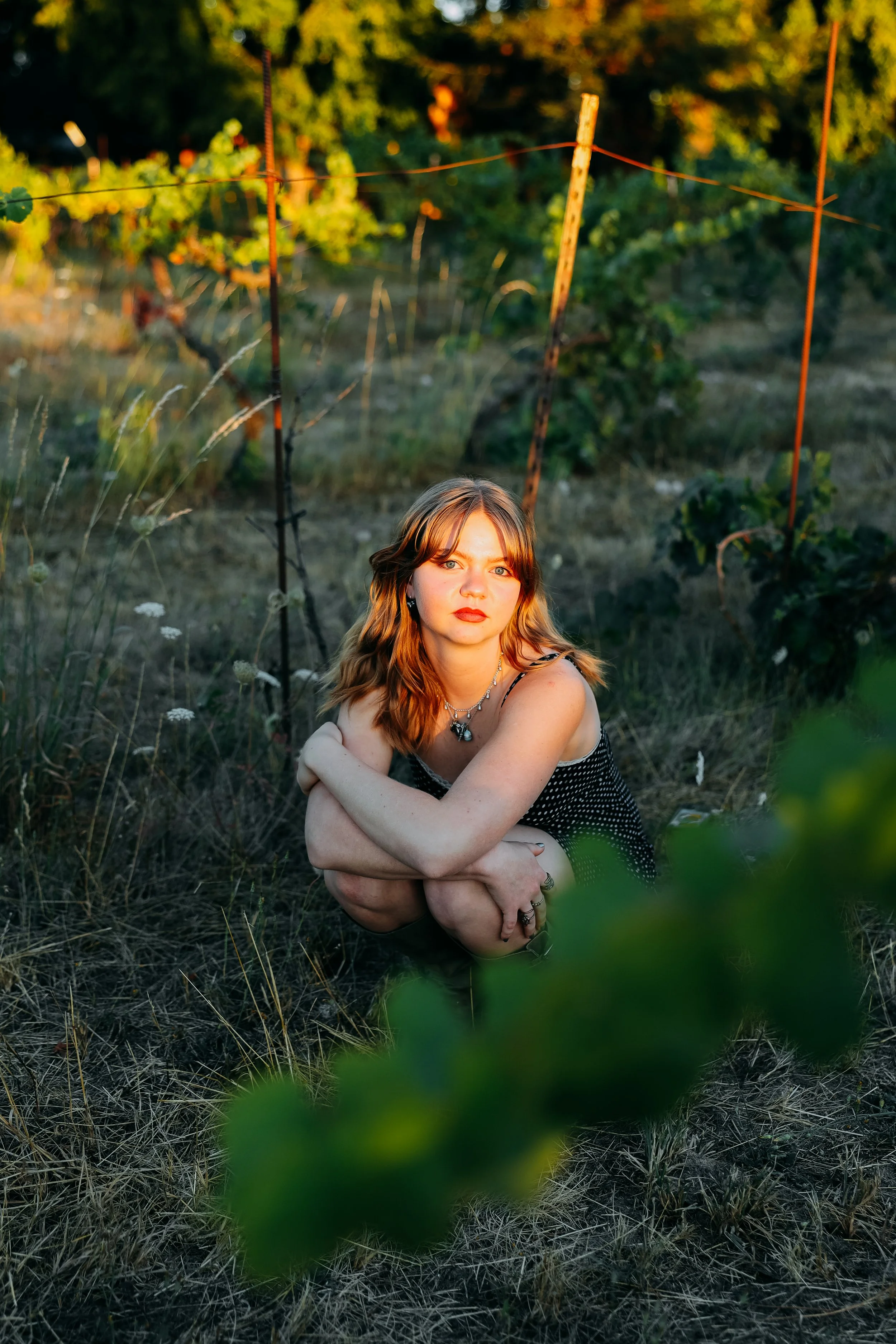 A young woman with shoulder-length red hair, wearing a black and white polka dot dress and jewelry, crouches on dry grass in a vineyard during golden hour, surrounded by green vines and orange stakes.
