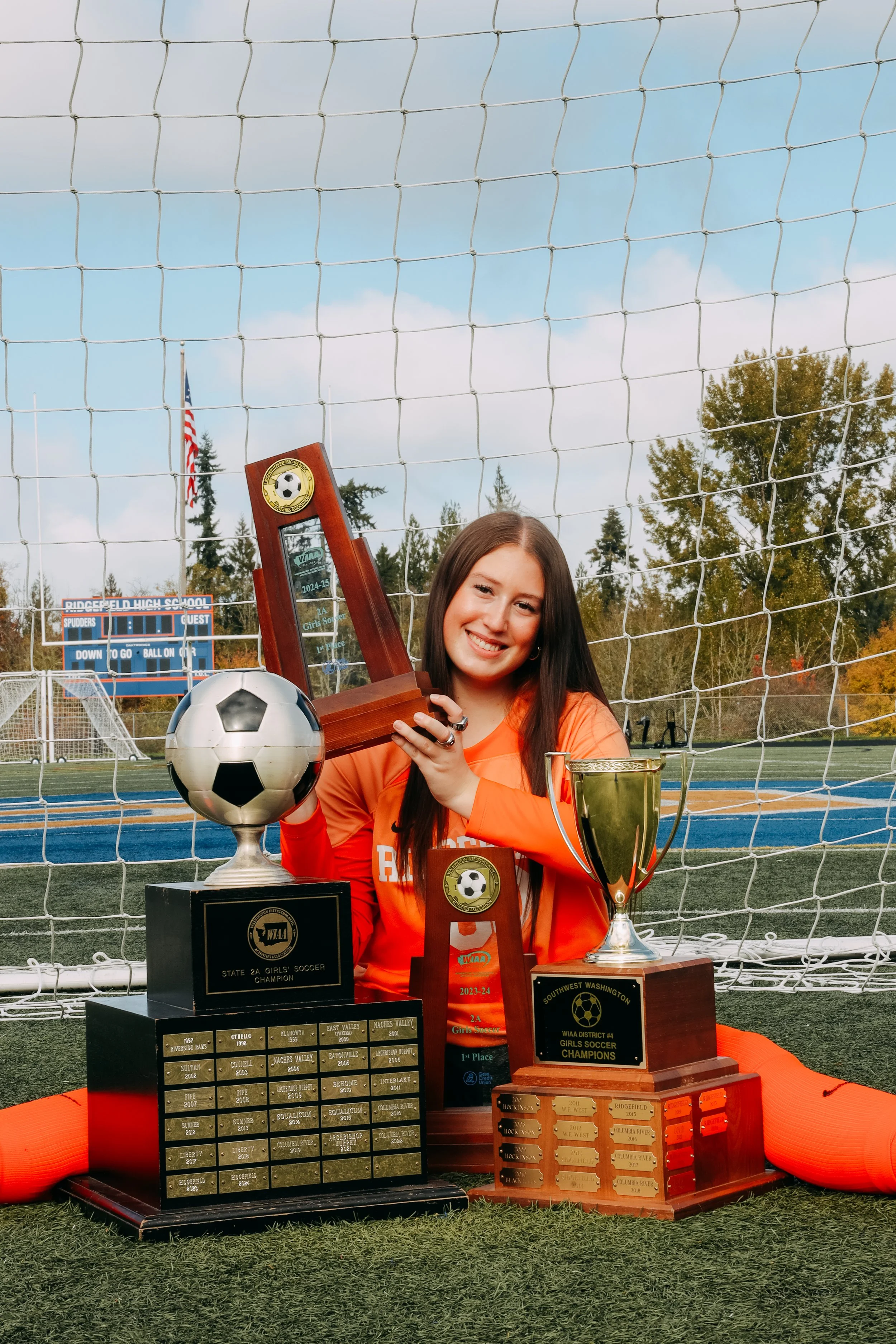 A young female soccer player sitting on a soccer field with several trophies and awards, holding her soccer championship trophy, smiling after a victory.