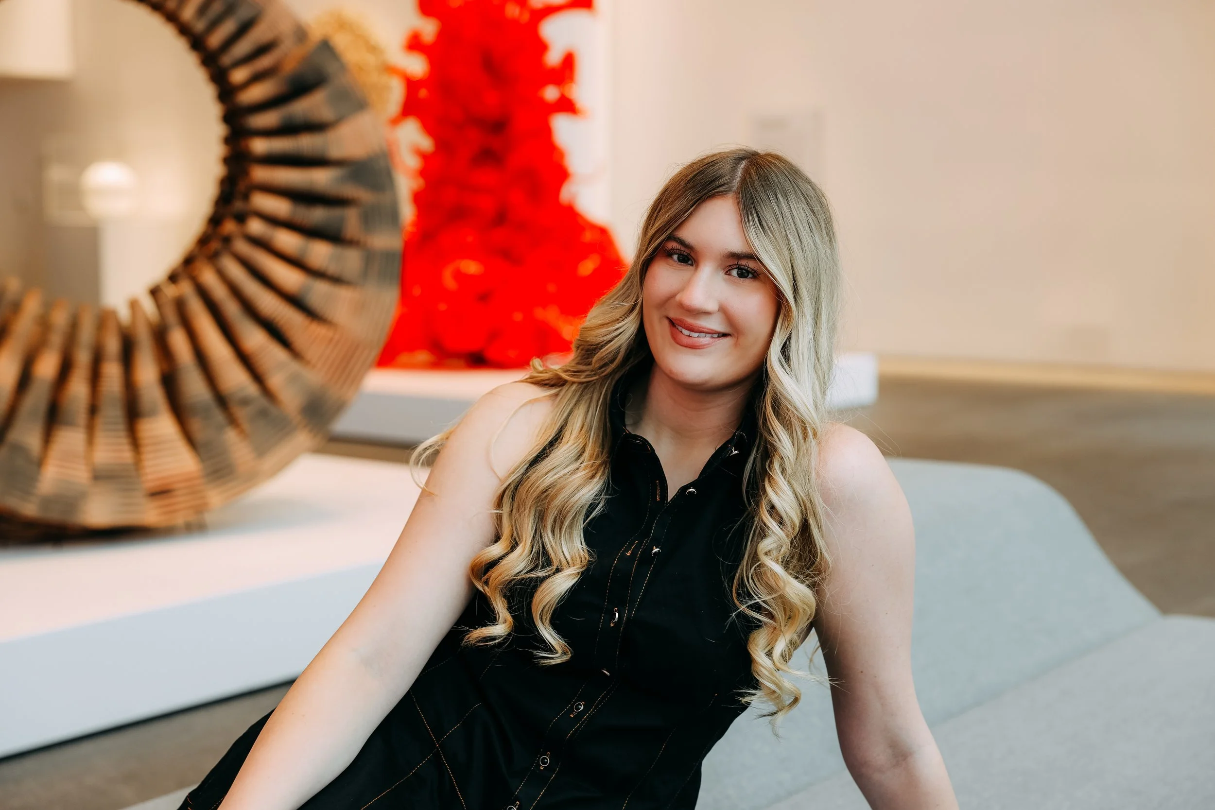 A young woman with long blonde hair smiling while sitting on a light gray sofa in a modern art gallery.