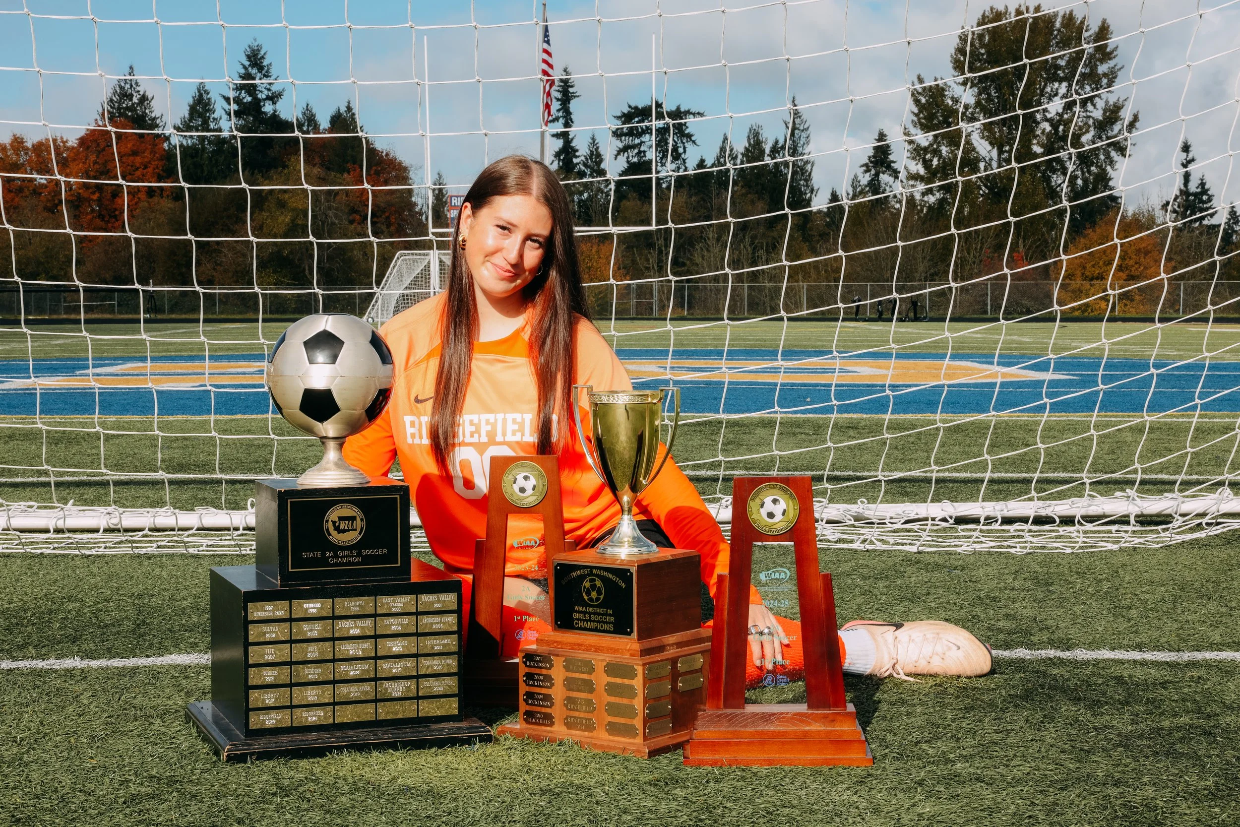 A young female soccer player sitting on a soccer field in front of a goal net, surrounded by trophies and awards, with trees and an American flag in the background.