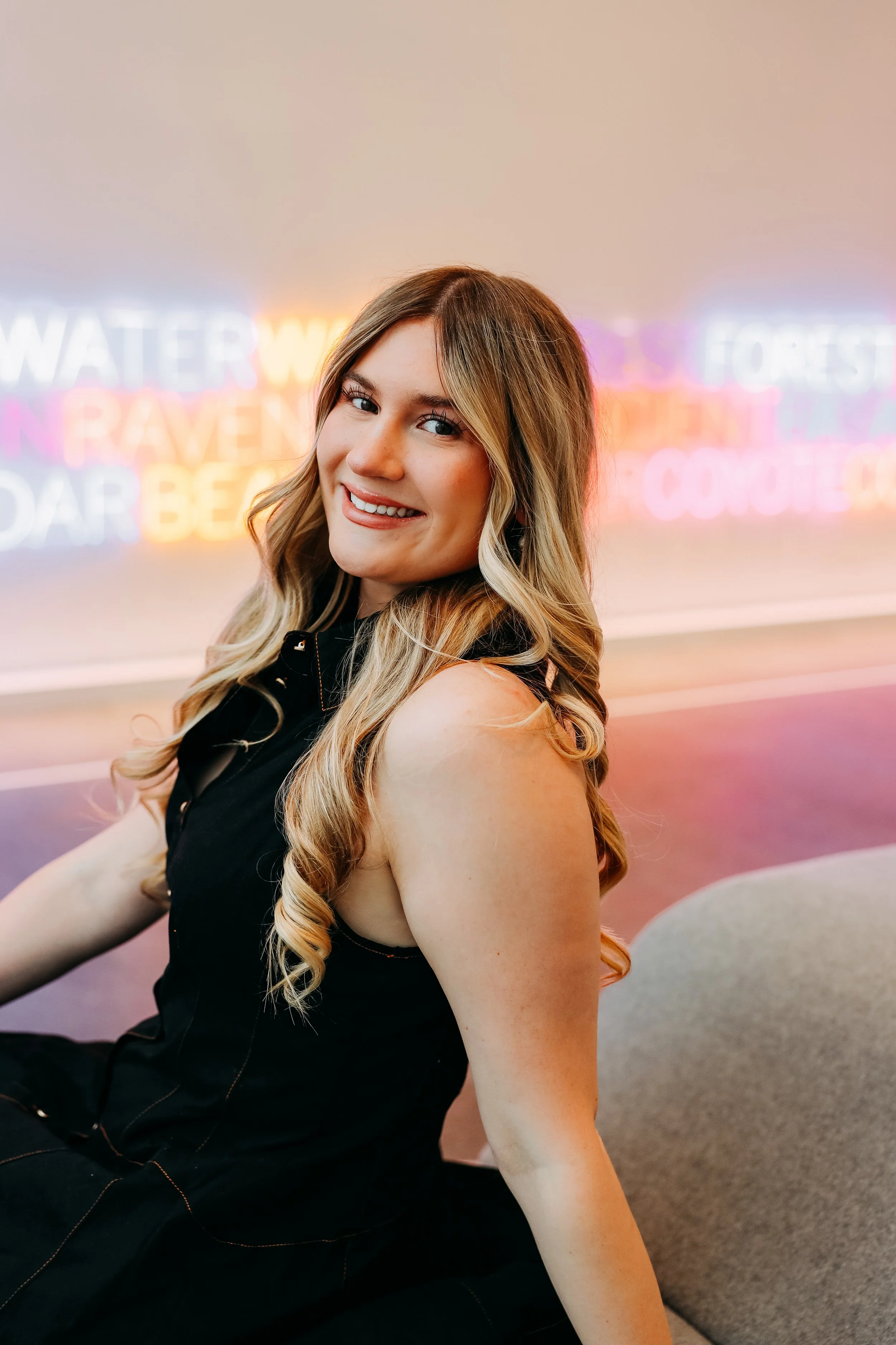 Portrait of a young woman with long, wavy blonde hair, smiling at the camera, sitting indoors with neon light signs in the background.
