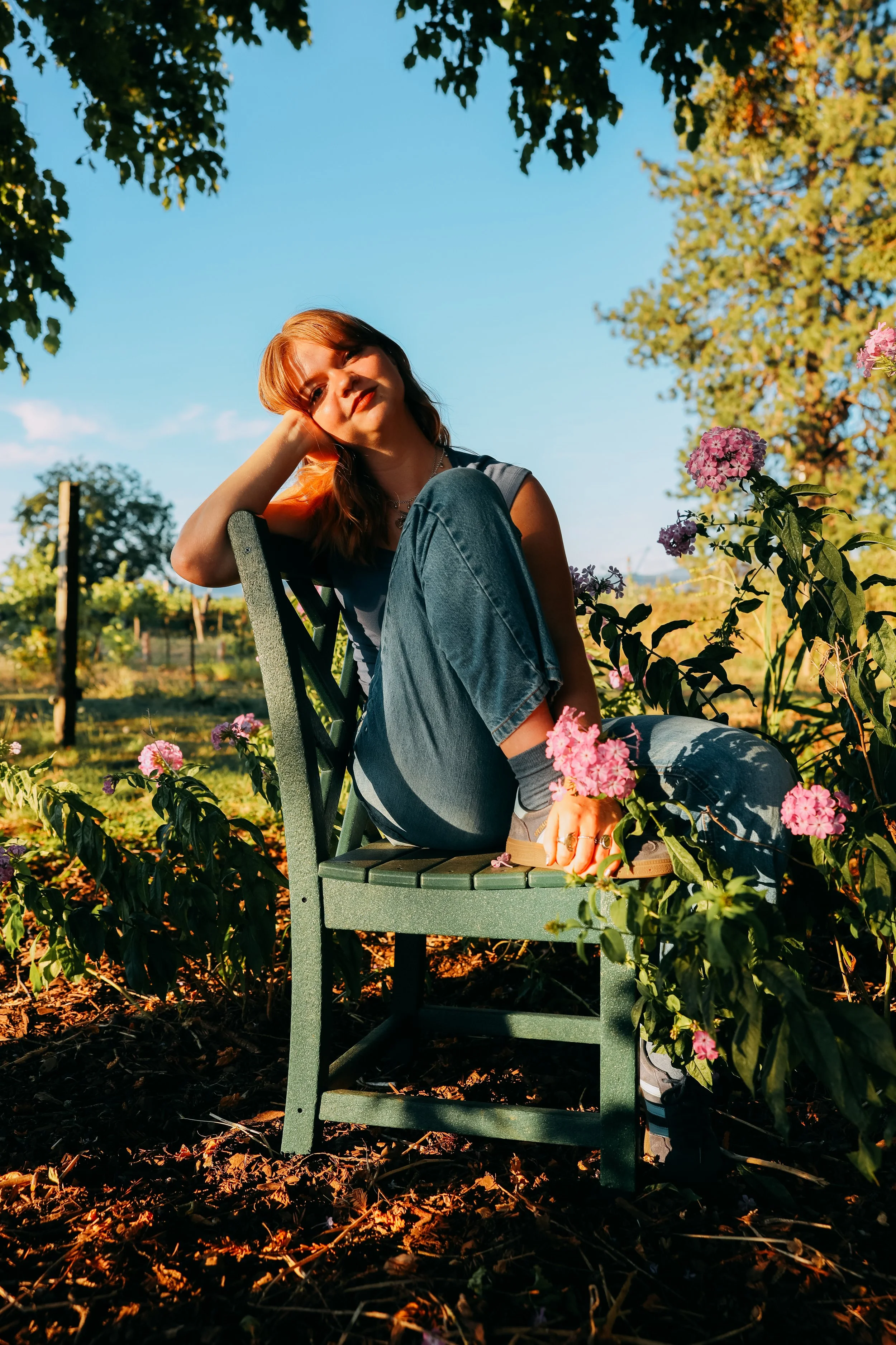 A woman with red hair sitting on a green garden bench surrounded by pink flowers in a garden during late afternoon. She is smiling, relaxed, looking at the camera, wearing a sleeveless top and jeans, with sunlight illuminating her face.