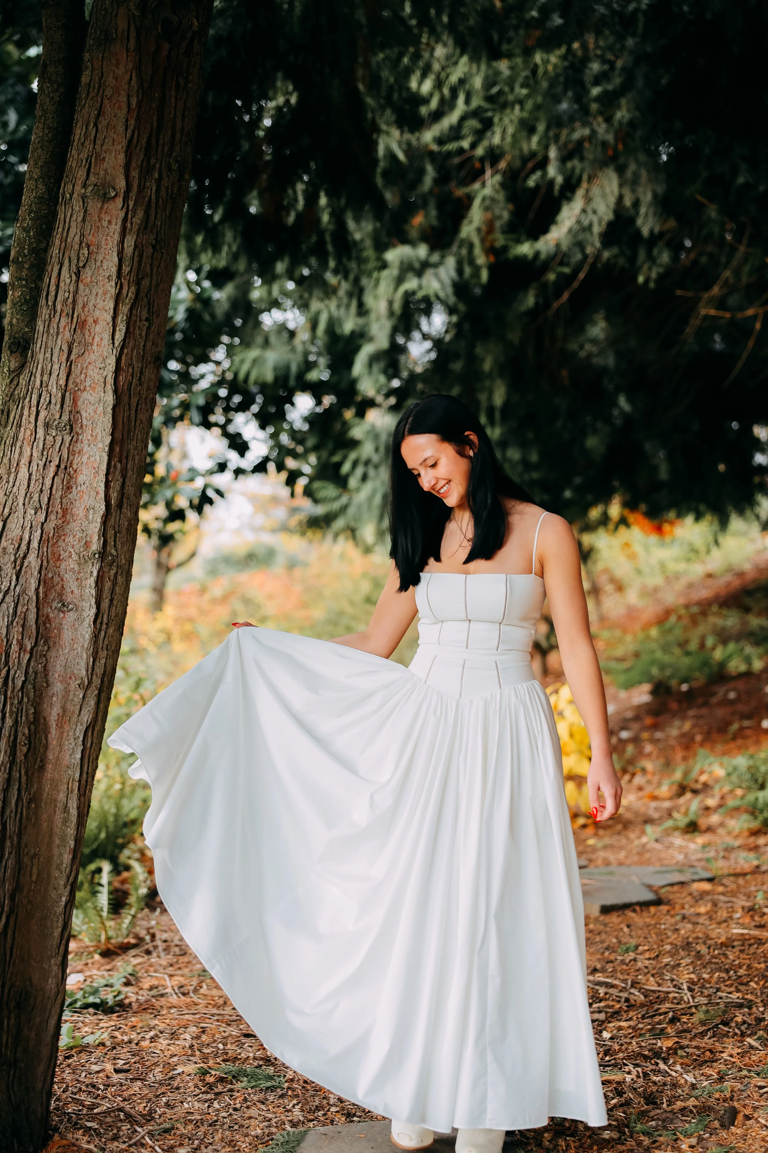 A woman in a white dress standing outdoors near a tree, smiling and holding her dress.