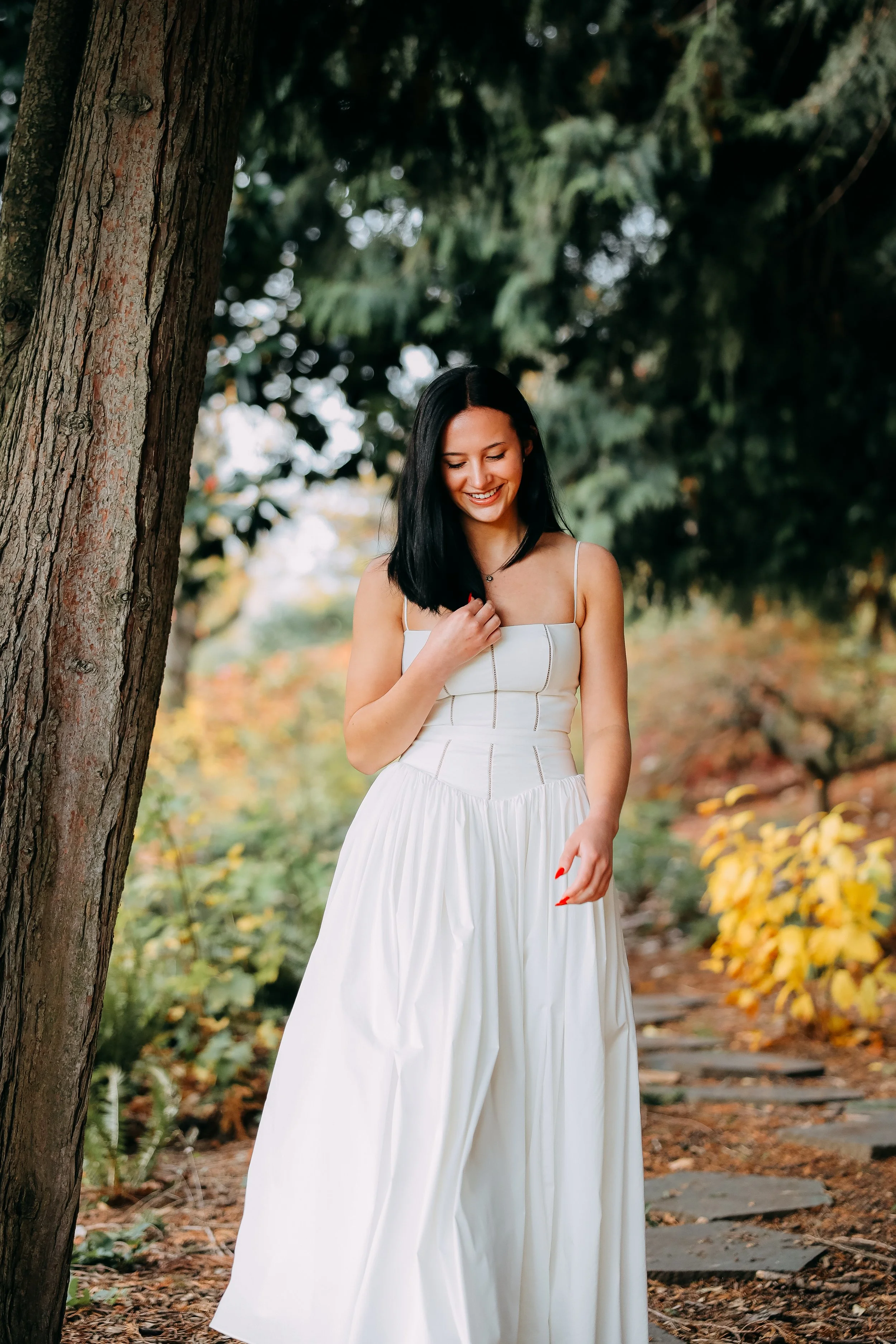 A woman in a white dress walking outdoors on a leaf-covered path surrounded by trees and yellow foliage.