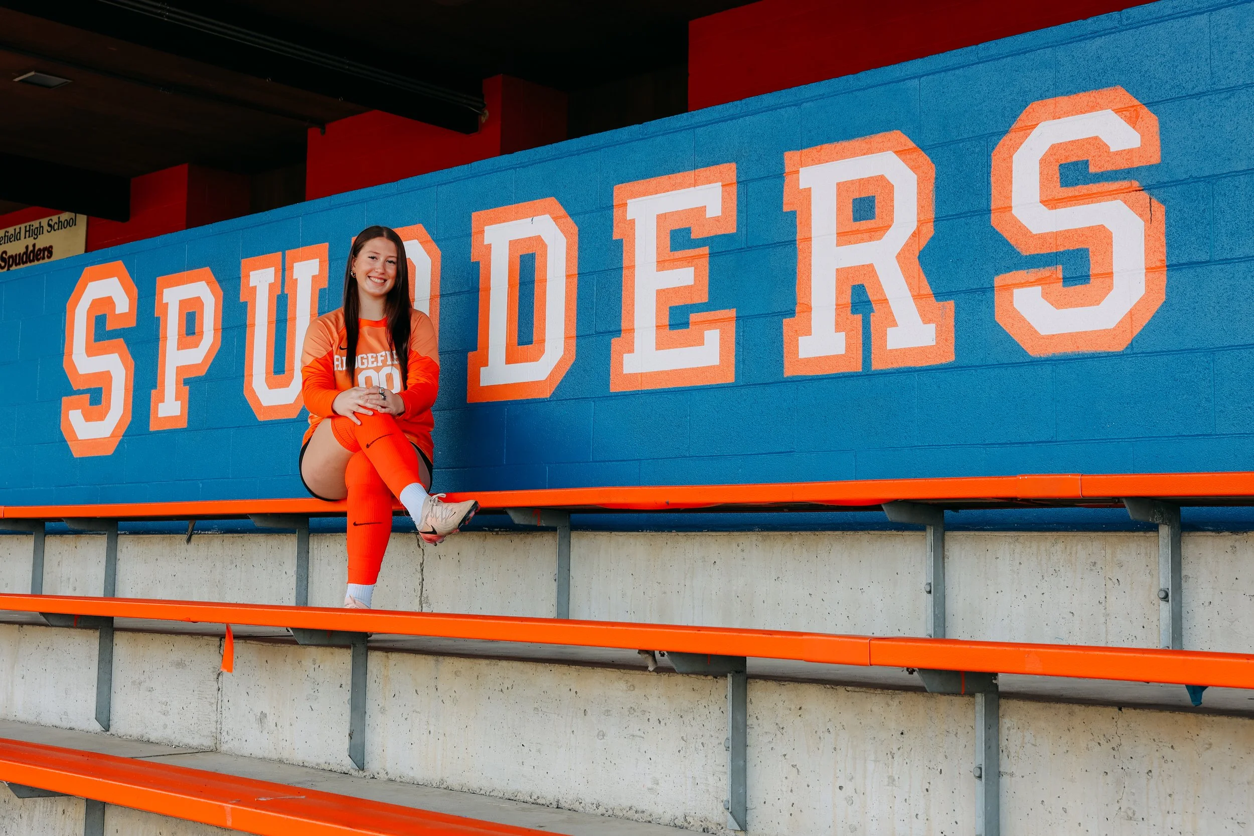 A young woman in sports uniform sitting on orange bleacher seat in front of a blue wall with the word SPUDERS written in large white and orange lettering.